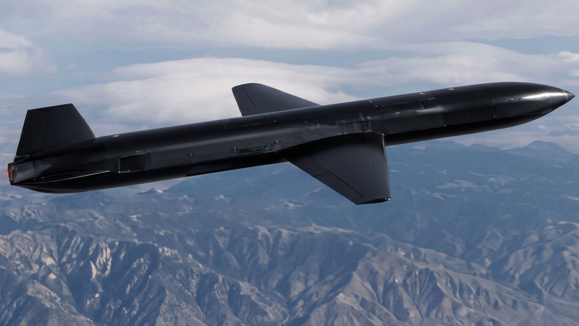 Dark, sleek unmanned aircraft with a long, cylindrical fuselage, mid-mounted small swept wings and a tall vertical tail, gliding over a jagged mountain range beneath a partly cloudy blue sky.