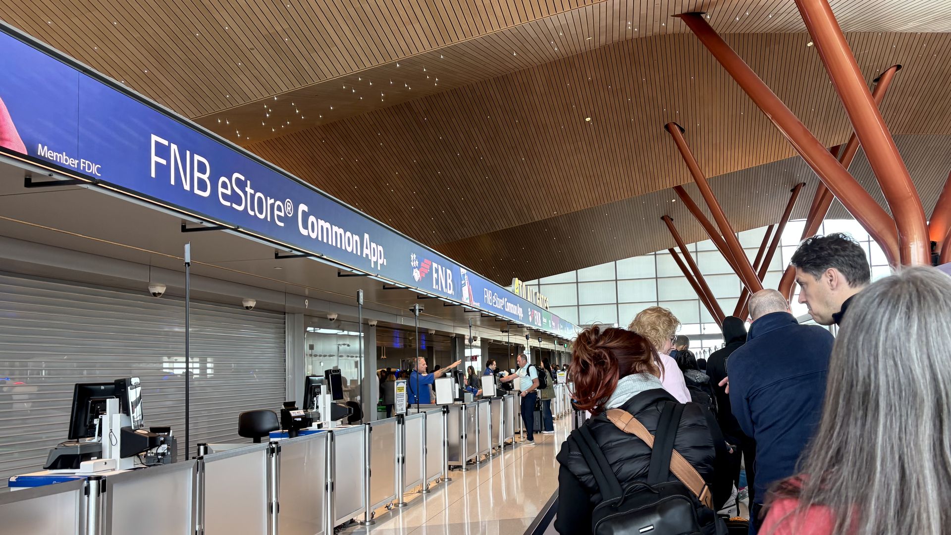 Inside an airport check-in hall, a blue sign reads FNB eStore Common App above glass counters. Orange curved supports rise over a wooden slat ceiling as passengers queue.