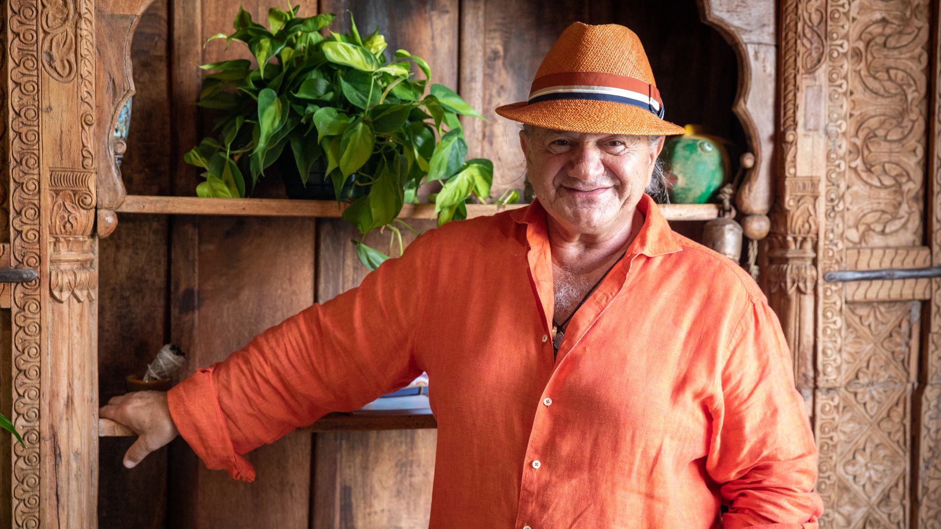 Giorgios Bakatsias stands wearing an orange shirt and a straw hat with a blue, white and red ribbon tied around it. 