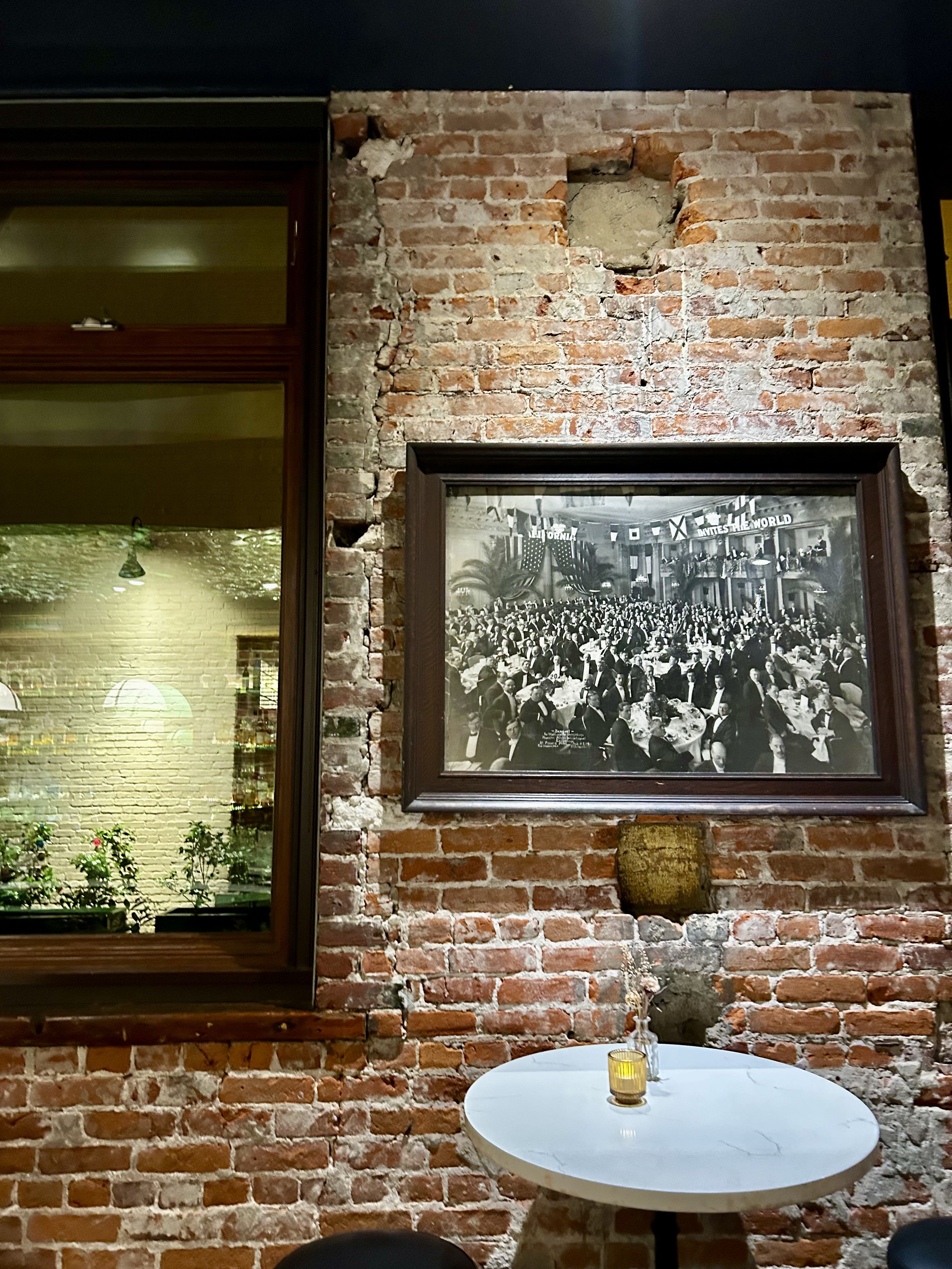 Brick wall with a dark wooden frame photograph of a crowded black-and-white banquet, hung above a small white round table with a lit candle; a wooden-framed window to the left shows a cozy interior.