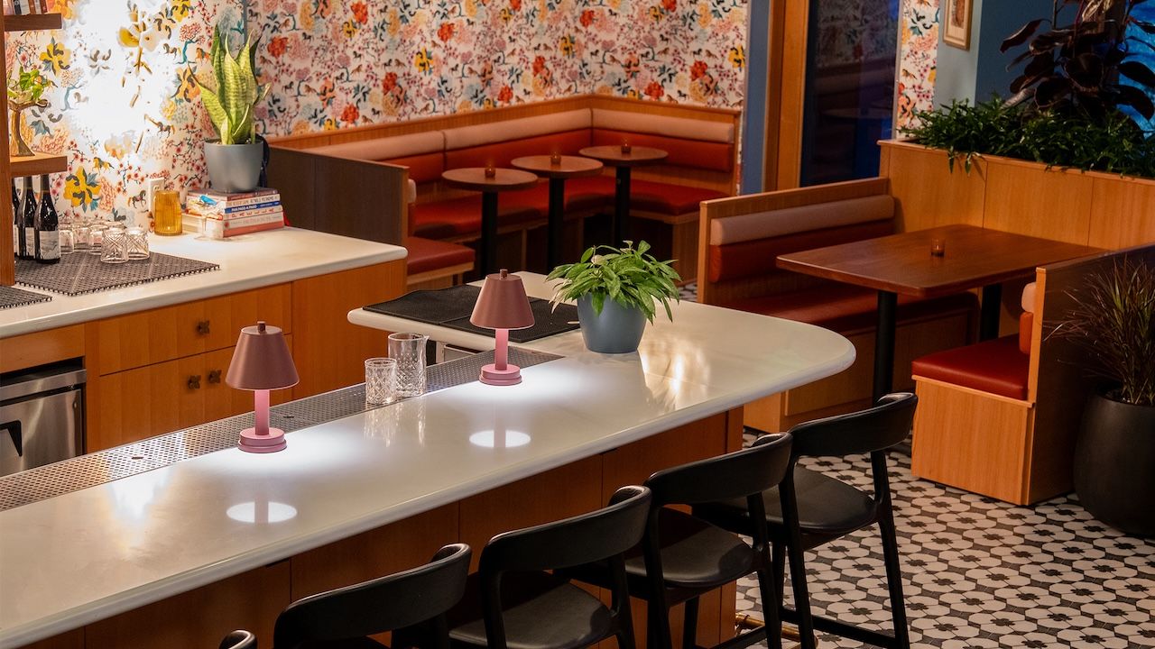 Bright retro cafe interior with a long white countertop, two pink lamps, and a potted plant. Floral wallpaper, red booth seating, wooden tables, and black chairs on a black-and-white tiled floor.