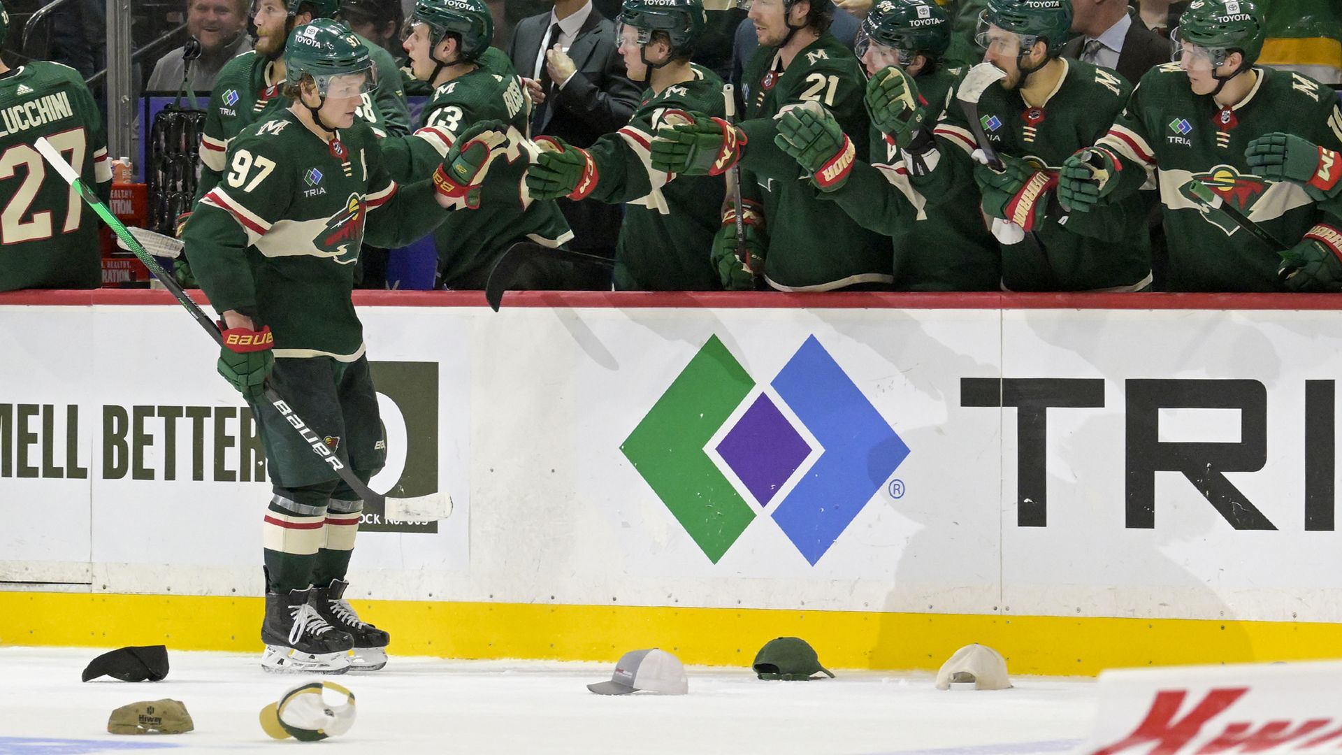 A hockey player in a dark green jersey with cream-colored trim skates along his team's bench, fist-bumping his teammates over the boards. Hats from the player's "hat trick" third goal of the game litter the ice.