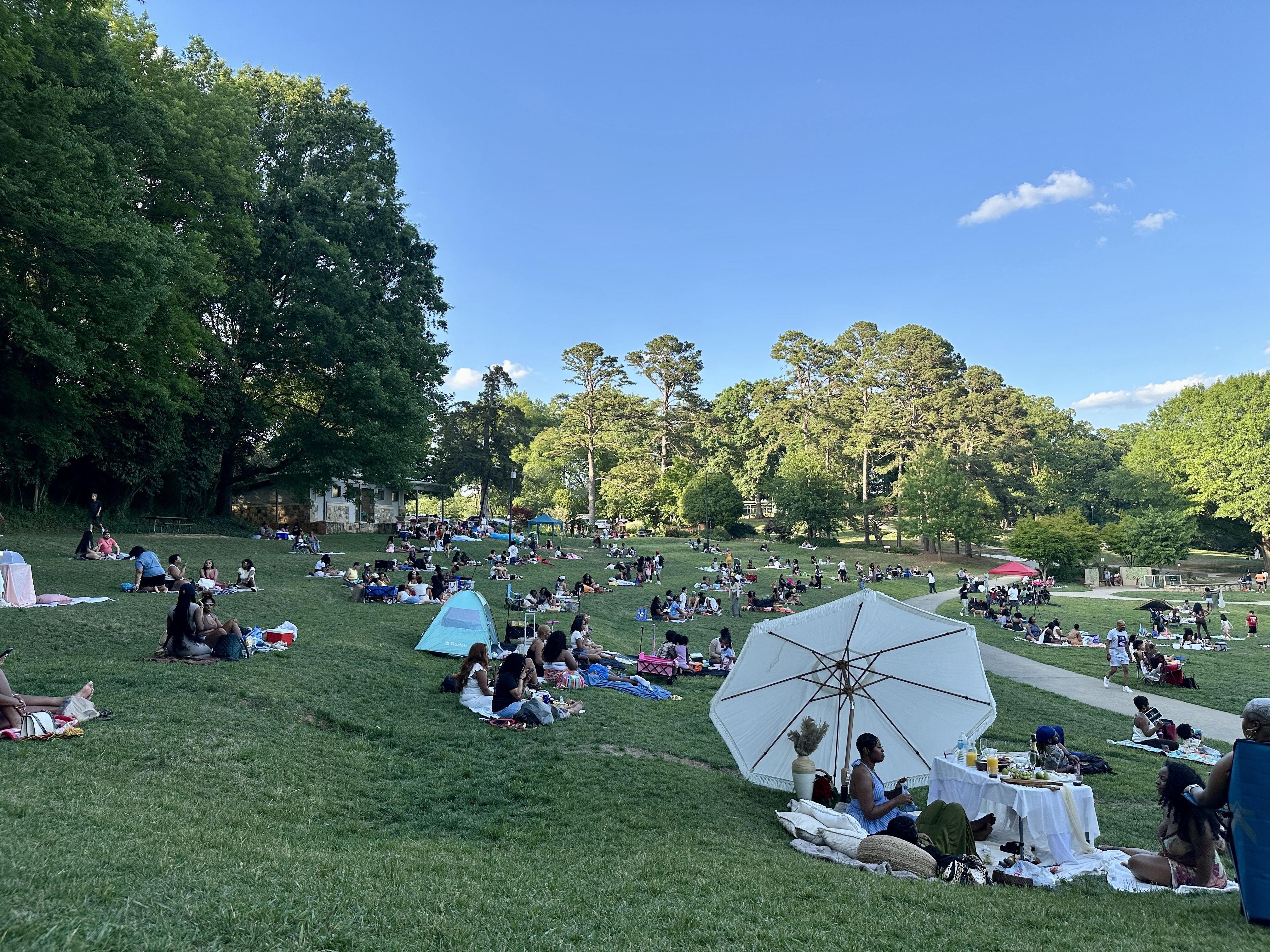 Sunny park with many families picnicking on a grassy lawn near trees. Tents and a white canopy are in the foreground; groups sit on blankets under a clear blue sky with a few clouds.