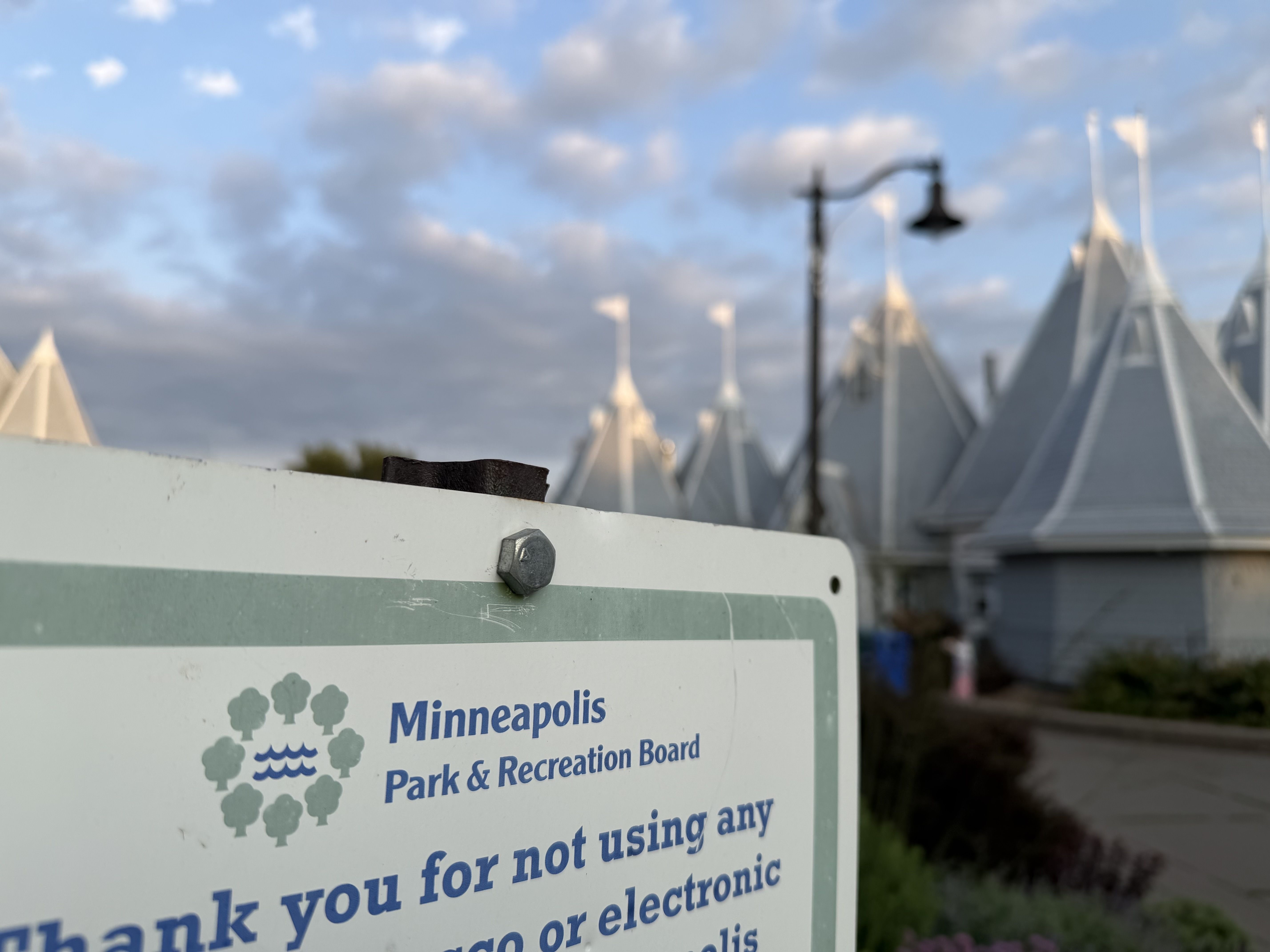 Close-up of a sign that says Minneapolis Park & Recreation Board with a blurred background of white tent shapes, a black street lamp, and a blue sky with clouds.