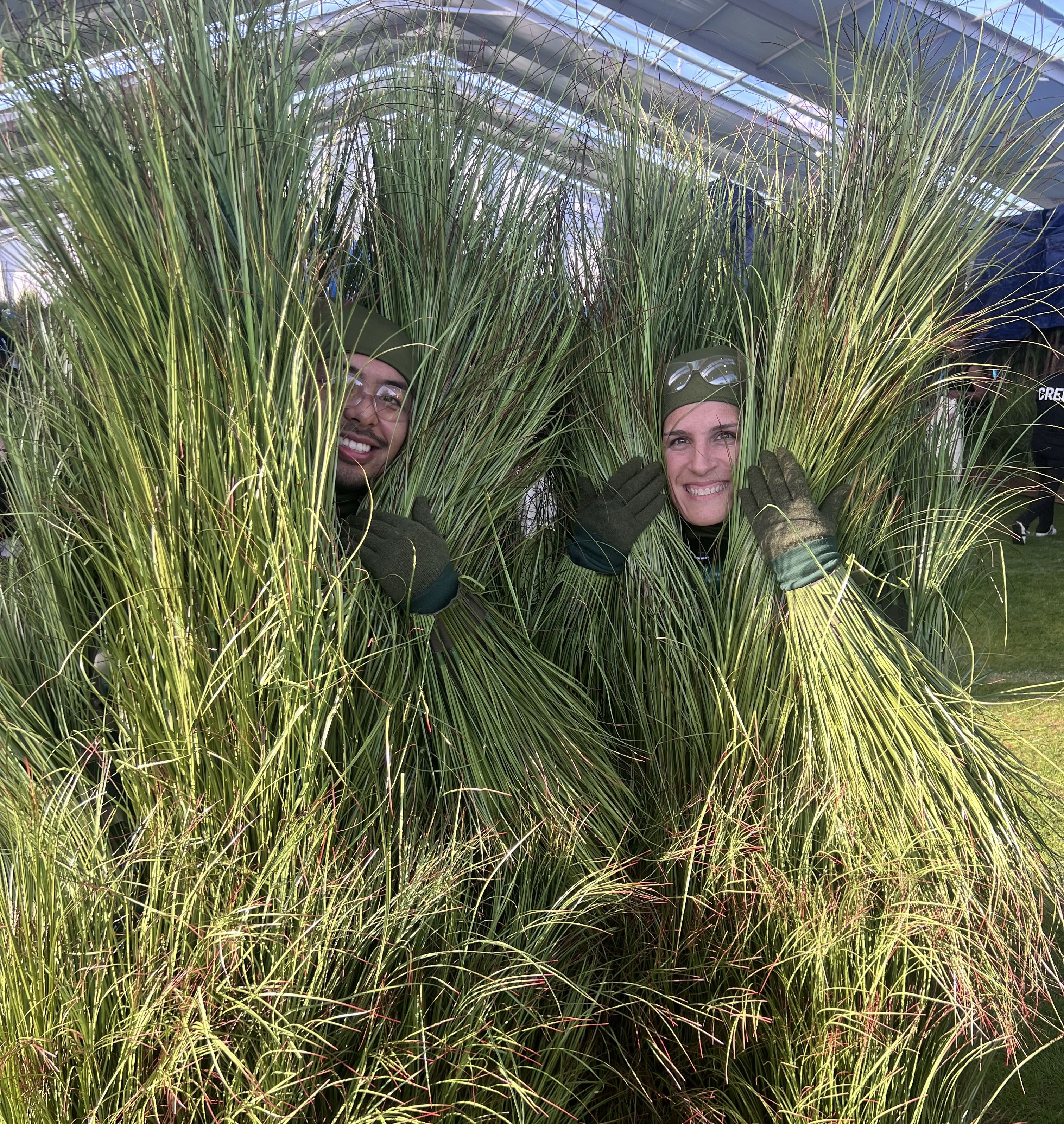 Two people peek out from behind costumes made of plastic grass, while wearing grass gloves, in a backstage tent on a sunny day.