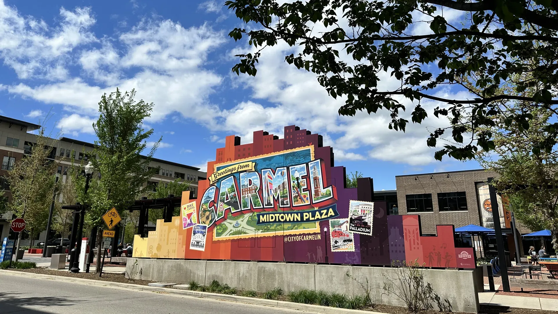 Colorful sign reading "Greetings from Carmel Midtown Plaza" with large block letters and postcard imagery, set against a blue sky with white clouds; tree branches frame the scene with buildings in the background.
