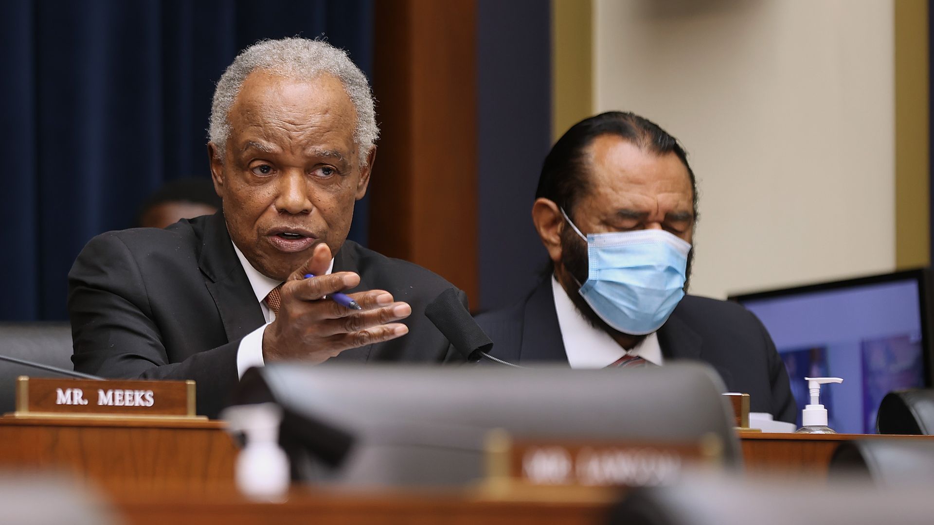 Two men at a formal hearing; left, older man with gray hair speaks and gestures with a pen, while right, a man wearing a blue surgical mask listens. A nameplate reads MR. MEEKS.