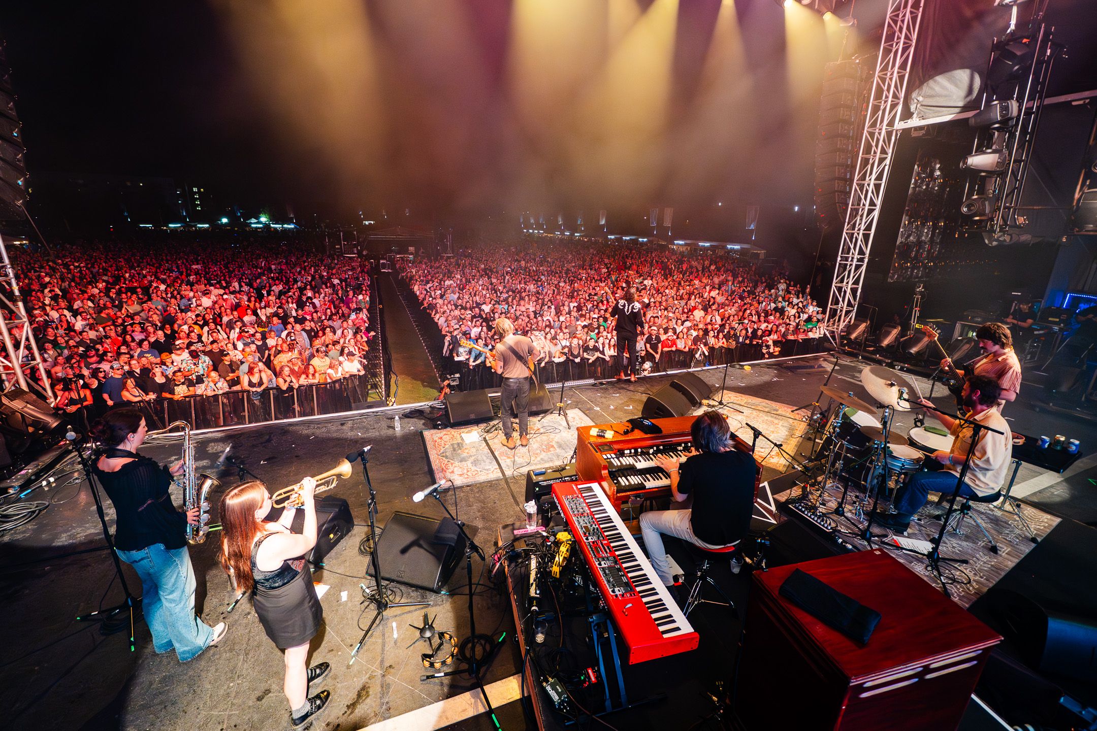 A crowded outdoor concert viewed from the stage: musicians play keyboards and drums as a dense audience watches. Yellow stage lights wash the crowd, with tall rigging and speakers on the sides.