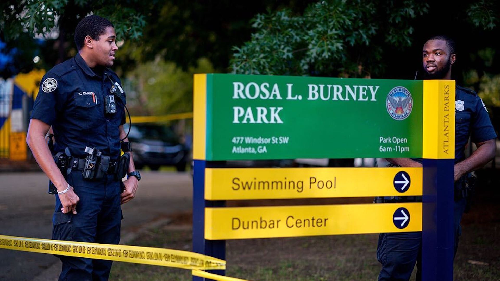 police officers standing next to park entry sign with crime tape wrapped around it