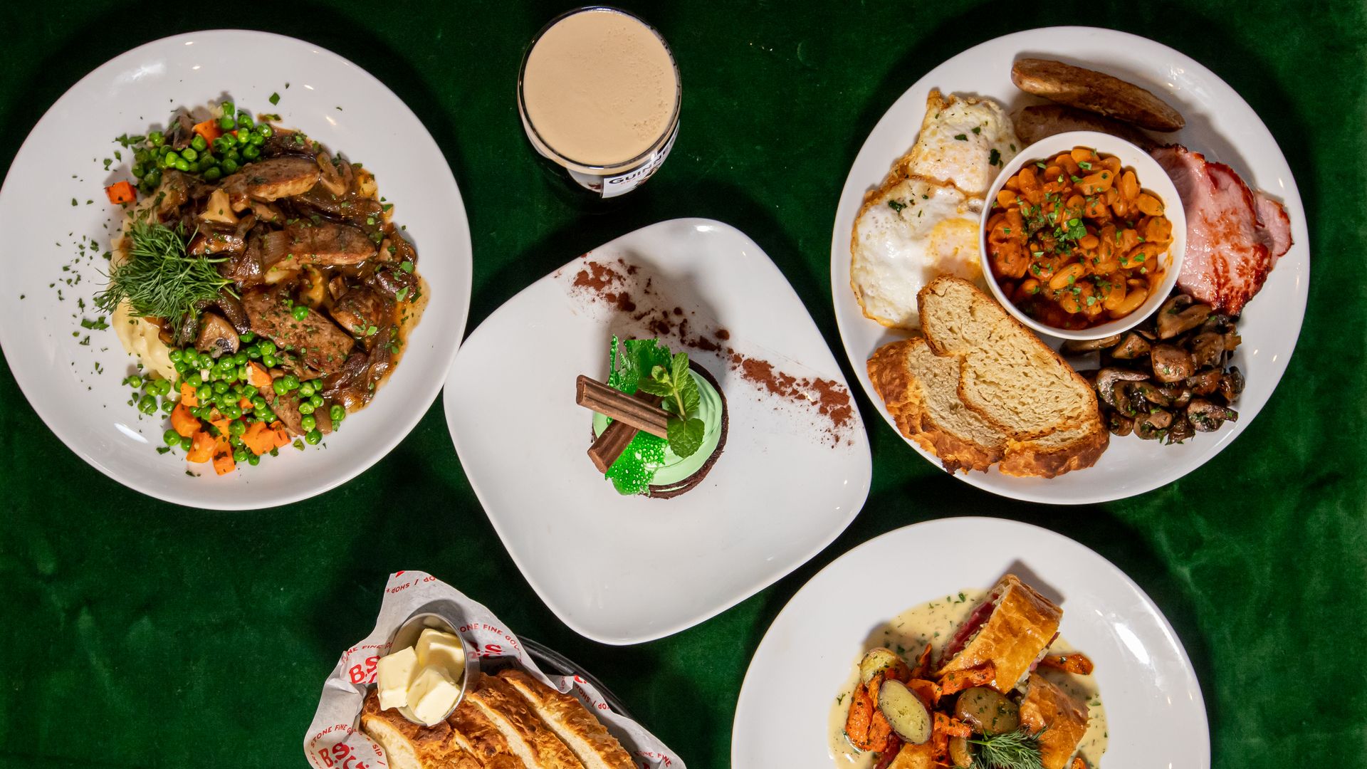 Top-down view of a green table with several plates: beef stew with peas and carrots; a green mint dessert with chocolate sticks; a breakfast plate with eggs and beans; bread with butter; and a veggie pastry.
