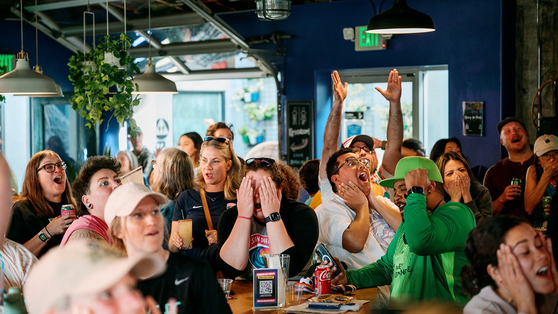 Crowded indoor bar with a cheering crowd watching a game. A man raises both arms in excitement while others clap or cover their faces; green/blue decor and hanging lights.