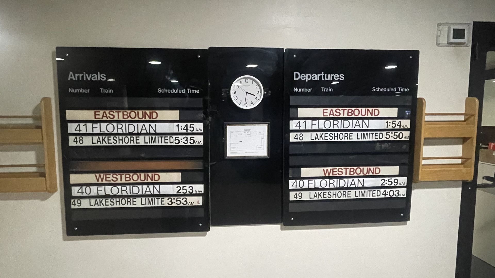 Black train timetable boards labeled Arrivals and Departures flank a central clock; Eastbound and Westbound schedules show Floridian and Lakeshore Limited trains with times; wooden benches on both sides.