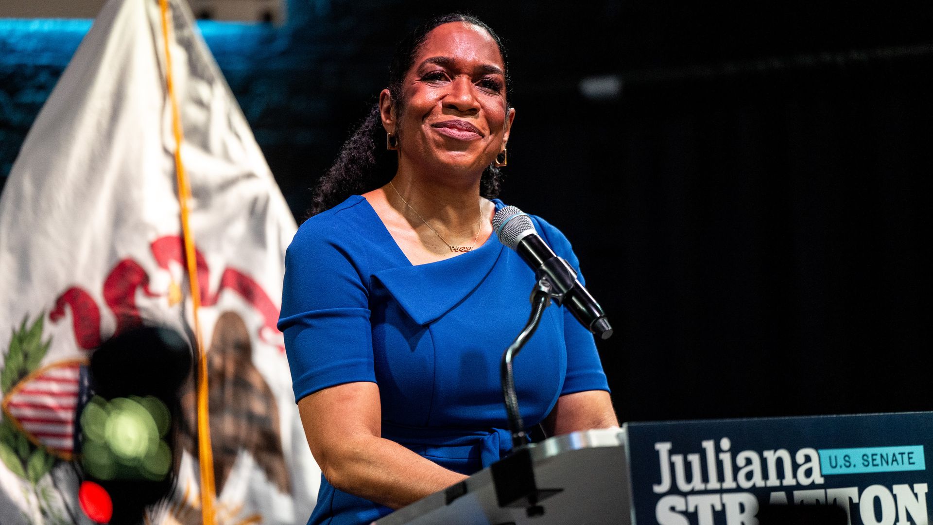 Smiling woman in a royal blue dress speaks at a podium with a microphone at a campaign event; the podium sign reads "Juliana Stanton" for U.S. Senate, with a colorful banner in the background.