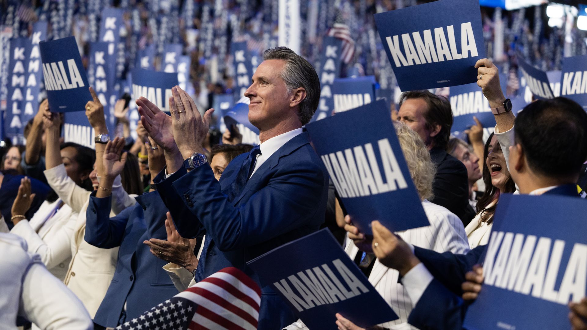 Gavin Newsom, wearing a dark blue suit, claps as people around him hold blue signs that say "KAMALA" in all-caps white letters.