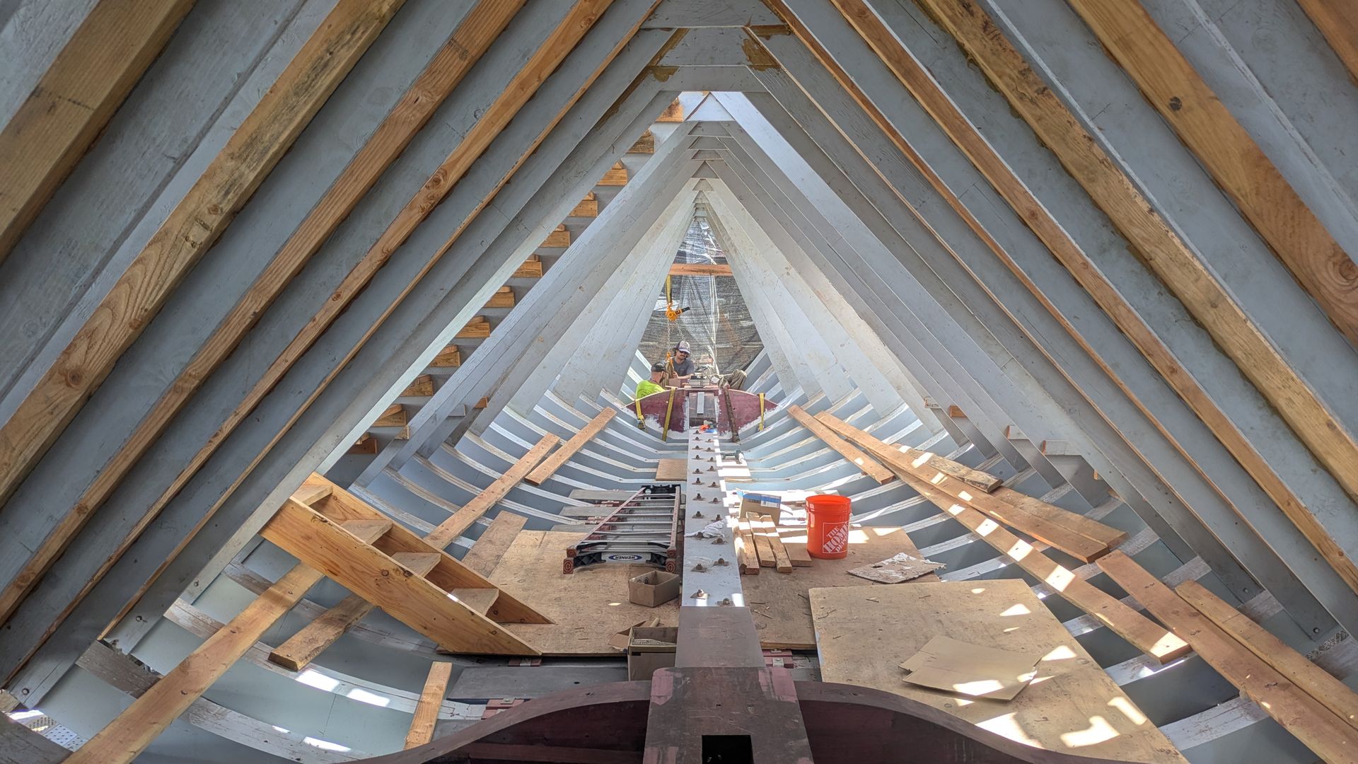 Inside a building under construction, a long row of blue-gray and natural wood roof trusses forms a tunnel-like perspective toward workers at the far end, with scattered planks and an orange bucket.