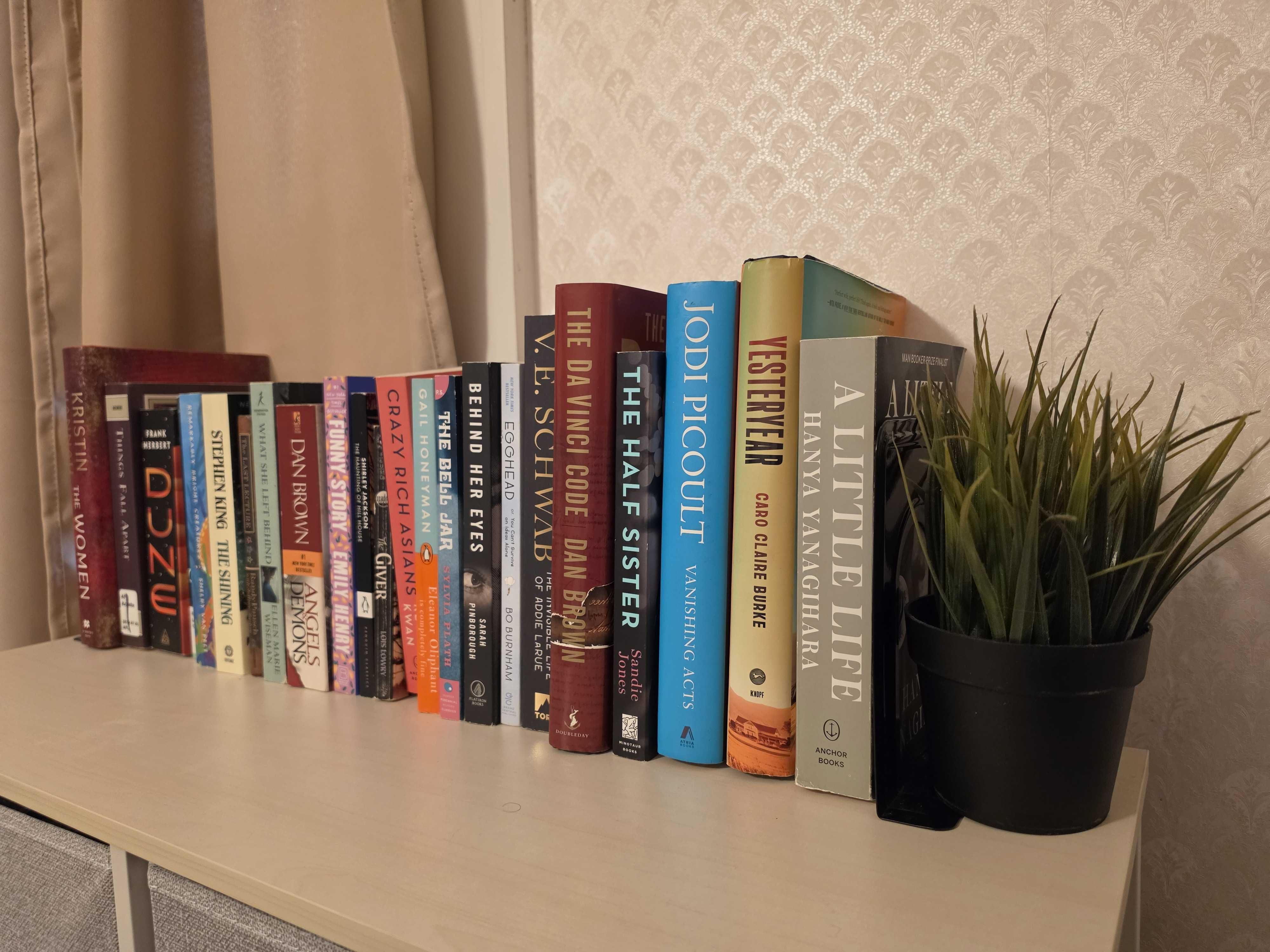 Row of colorful books on a low shelf beside a beige damask wall, with a beige curtain on the left and a potted plant on the right.