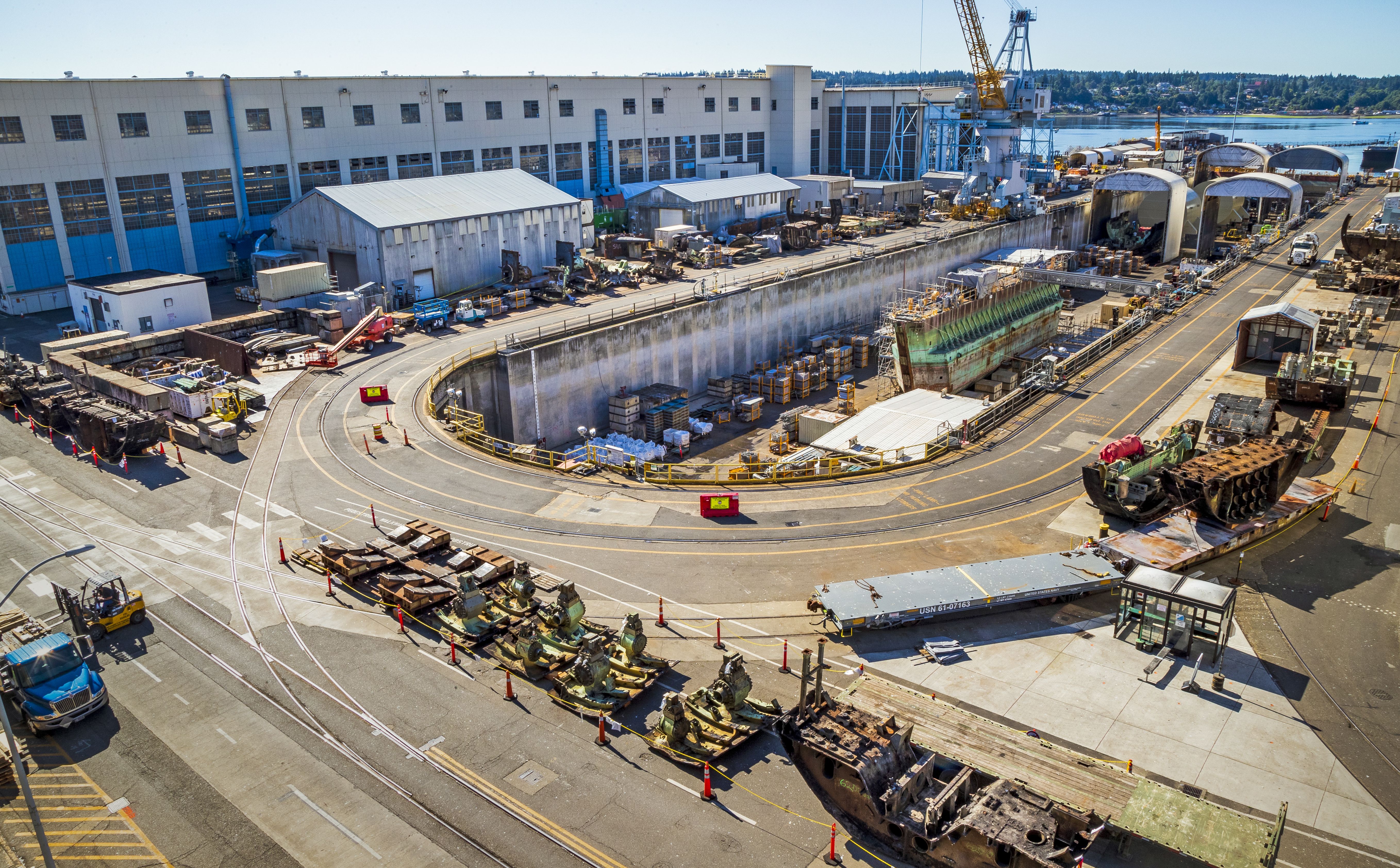 Aerial view of shipyard construction: white industrial buildings, a circular dry dock, cranes, crates, and scattered equipment along the waterfront.