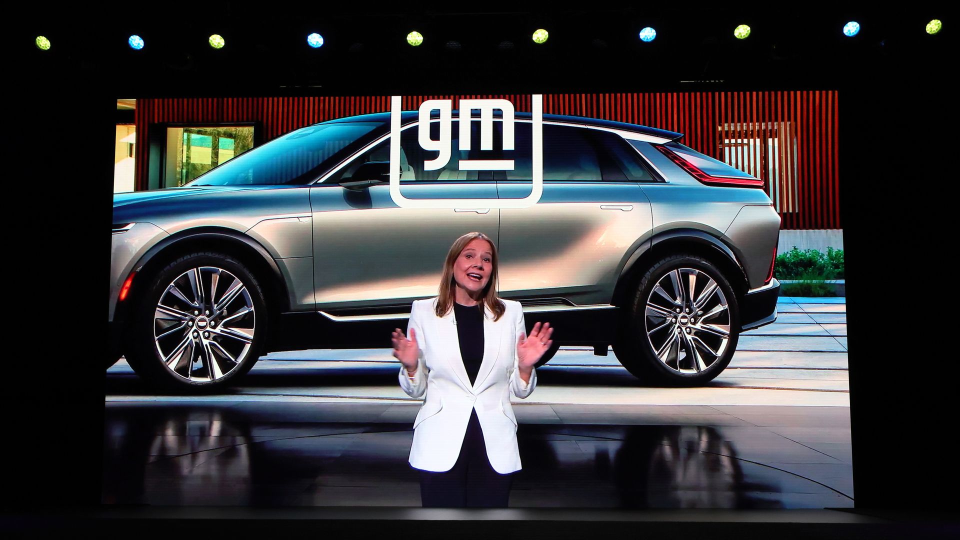 A woman in a white suit jacket stands with her hands spread open against a backdrop of an electronic screen with an SUV on it