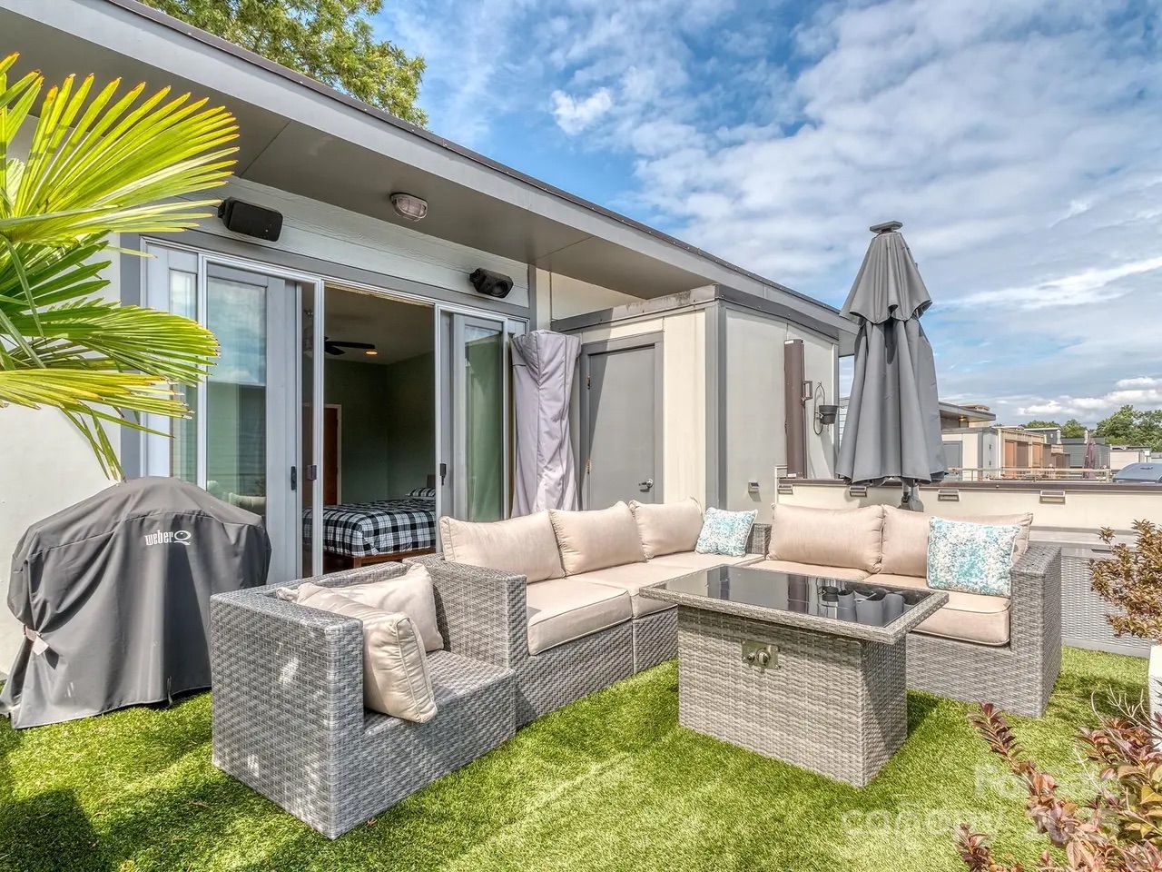 Outdoor patio with gray wicker L-shaped sofa with beige cushions, glass-top table, closed gray umbrella, grill covered with black Weber cover, and green artificial grass under blue sky.