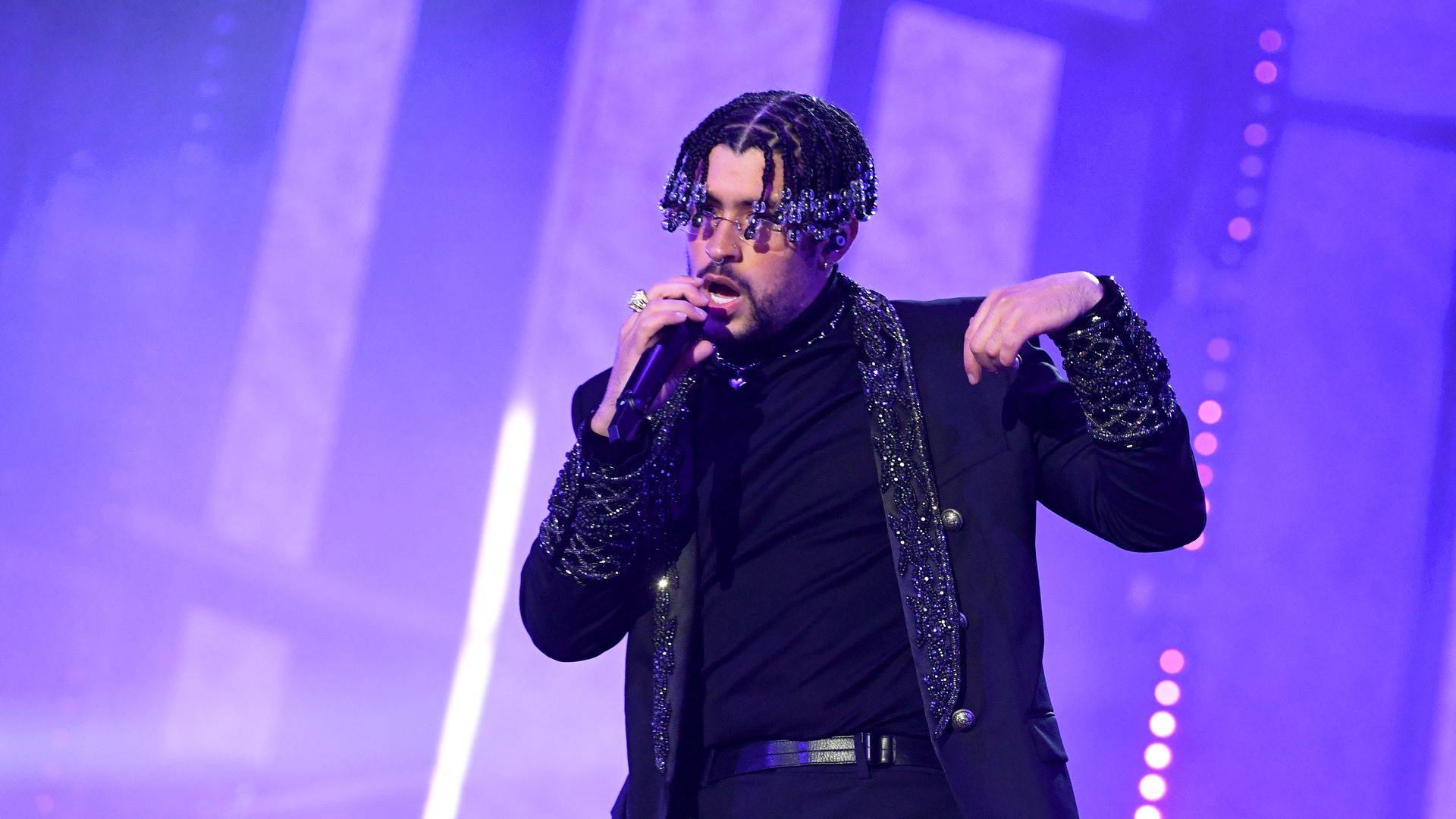 Bad Bunny performs on stage in a black sparkly blazer with purple lights behind him.
