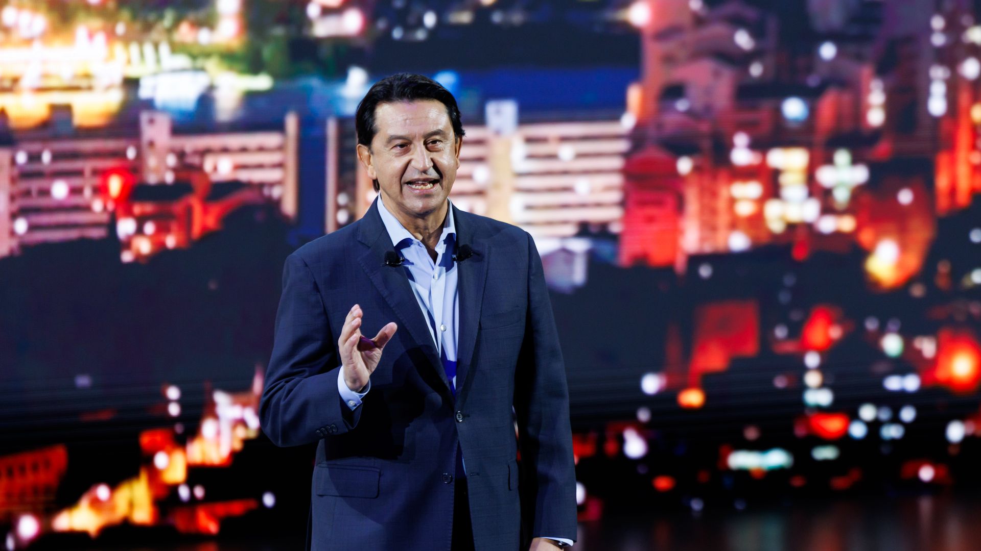 Hyundai Motor Co. CEO José Muñoz speaks in front of a colorful cityscape background while gesturing with his right hand. 