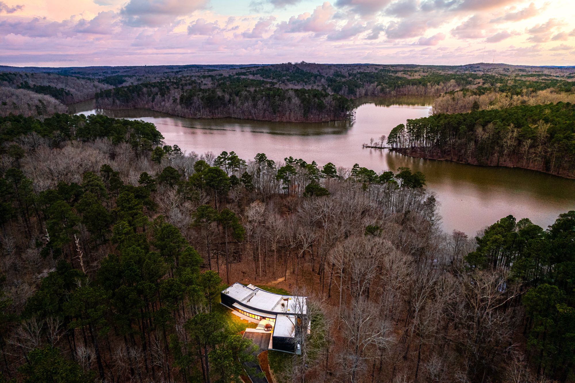A modern house with bright lights, surrounded by bare trees and green pines near a large winding lake under a pastel sunset sky.