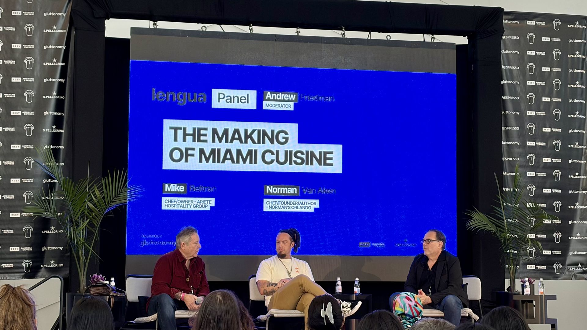 Panel discussion on a stage with a bright blue screen reading "The Making of Miami Cuisine." Three men seated in front of an audience; black branded banners, plants, and water bottles flank the setup.