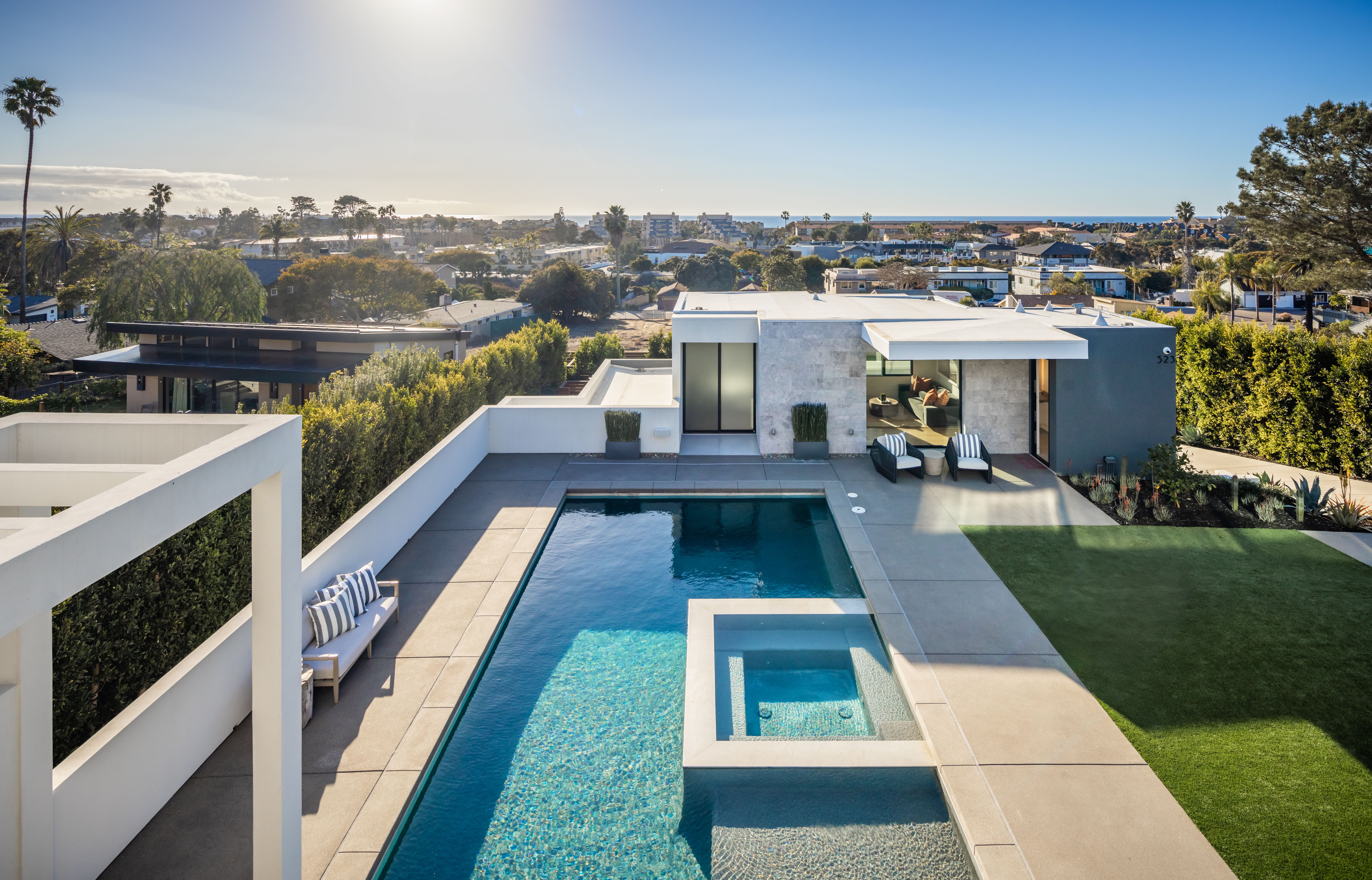 Modern house with a rectangular pool and lounge area in the foreground. Glass walls, stone accents, and a manicured lawn. A sunlit suburban neighborhood with palm trees stretches toward distant sea.
