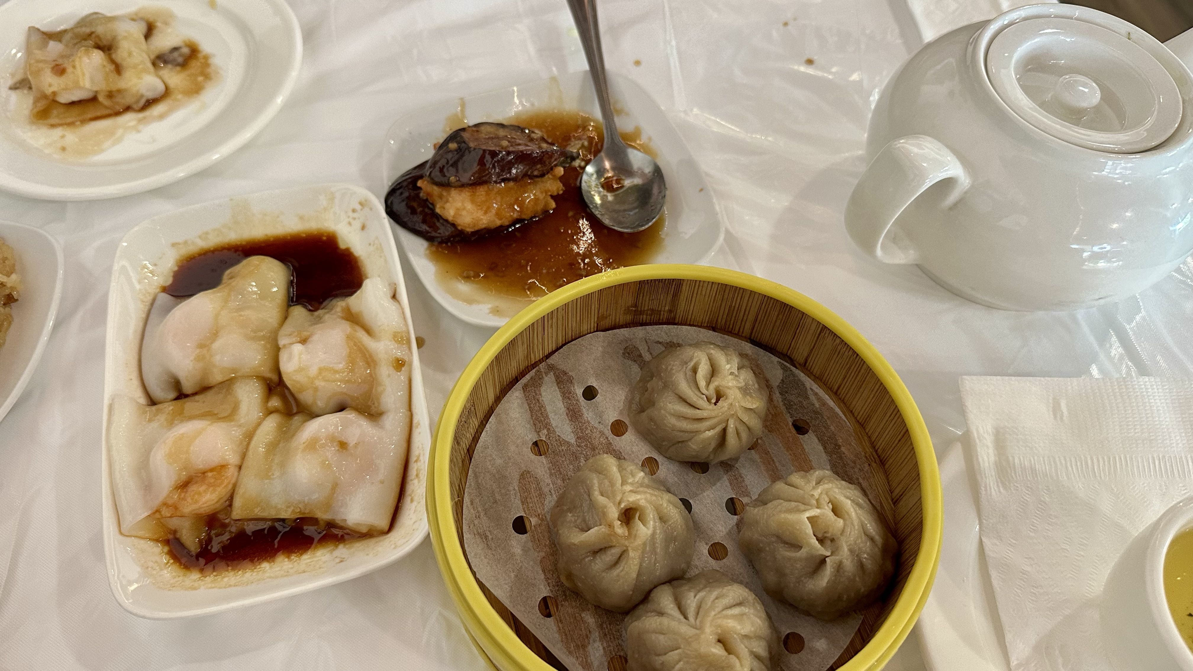 Overhead shot of a dim sum table: a yellow bamboo steamer with steamed buns, white plates of dumplings in soy sauce and braised meat, and a white teapot on a white table.