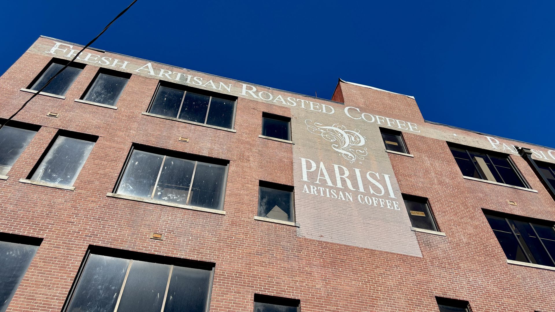 Red brick building with large windows and white painted sign reading "Fresh Artisan Roasted Coffee" and "Parisi Artisan Coffee" under bright blue sky.