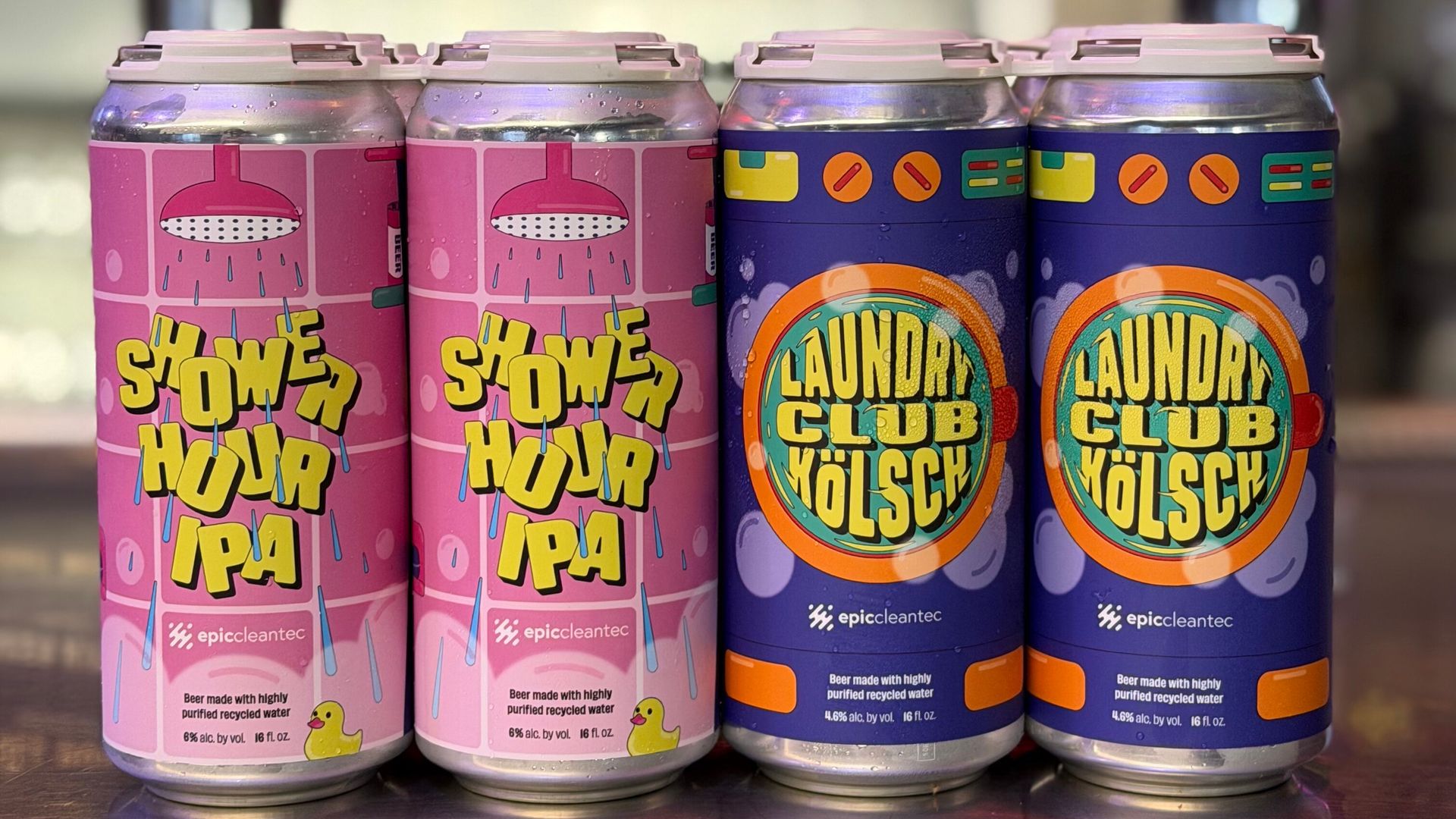 Two four-packs of beer cans on a reflective surface. Left pack is pink labeled "Shower Hour IPA" with showerhead and rubber duck design. Right pack is blue labeled "Laundry Club Kölsch" with washing machine dial design.