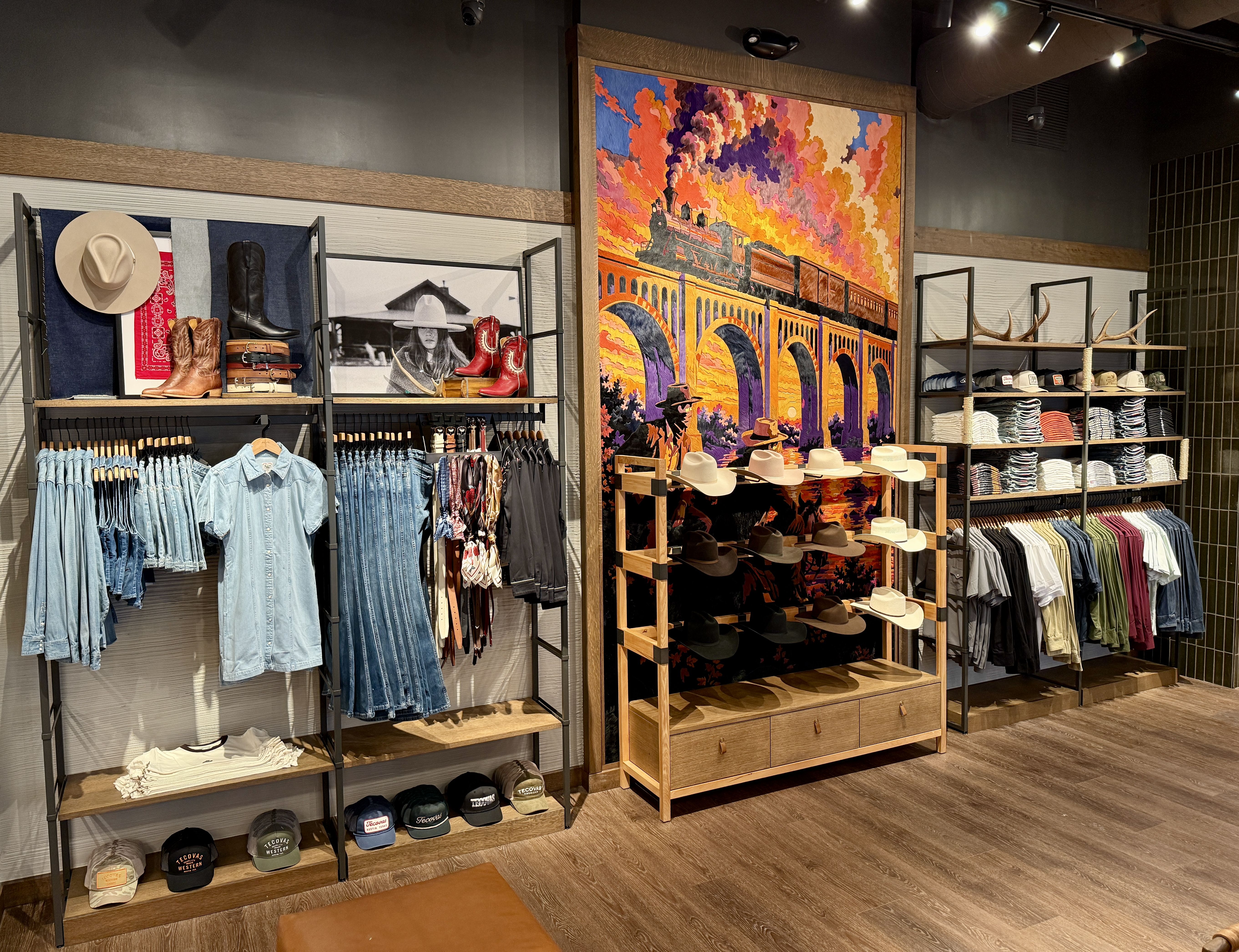 Inside a retail clothing store with racks of denim shirts, boots, hats, and belts; a large colorful mural of a train crossing an arched bridge dominates the back wall.