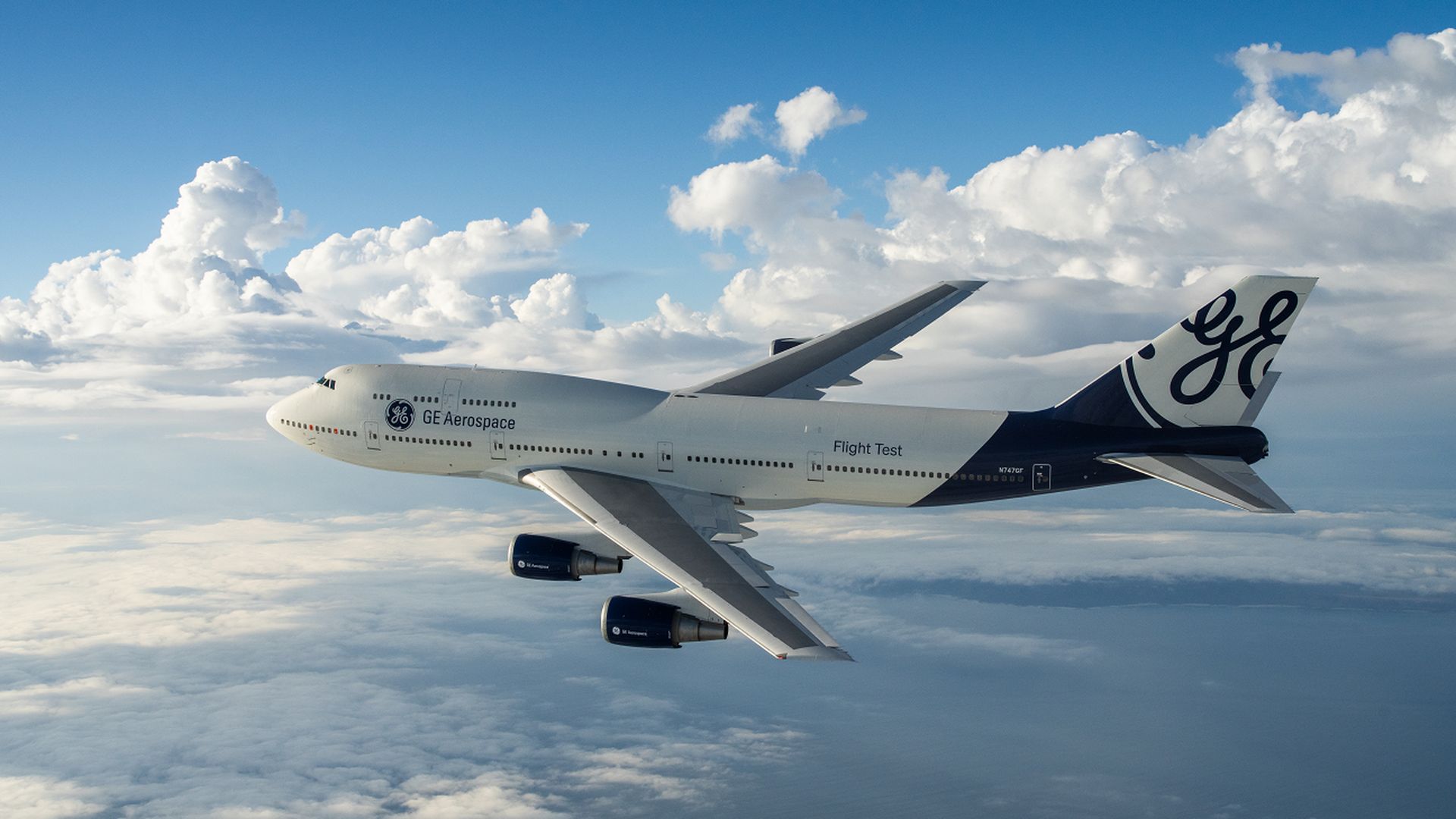 GE Aerospace wide-body aircraft in flight above clouds, four engines under the wings, white and blue livery with a large GE logo on the tail and Flight Test on the fuselage.