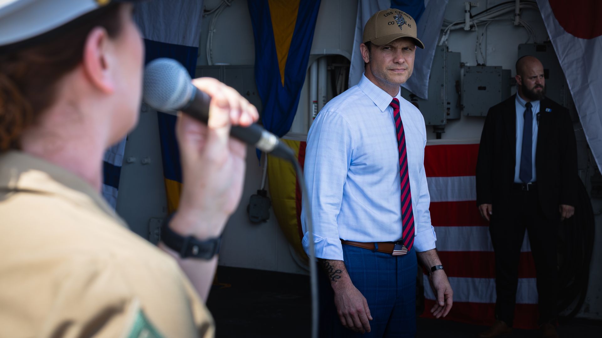 Pete Hegseth, a man, is seen watching a woman in naval military uniform sing into a microphone. Hegseth is wearing business casual and a brown ball cap.