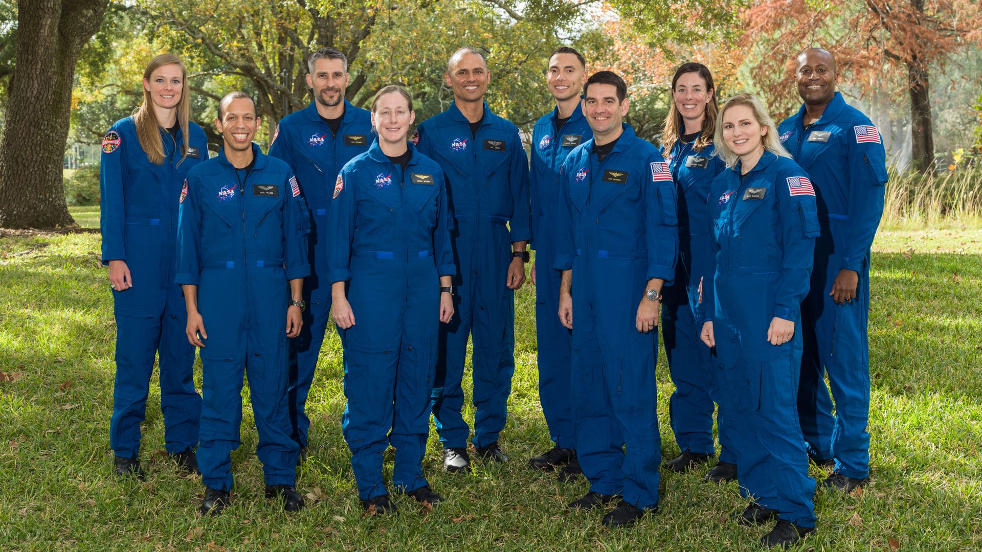 10 astronaut candidates in blue flight suits stand outside on a blue day under green trees. They're smiling.