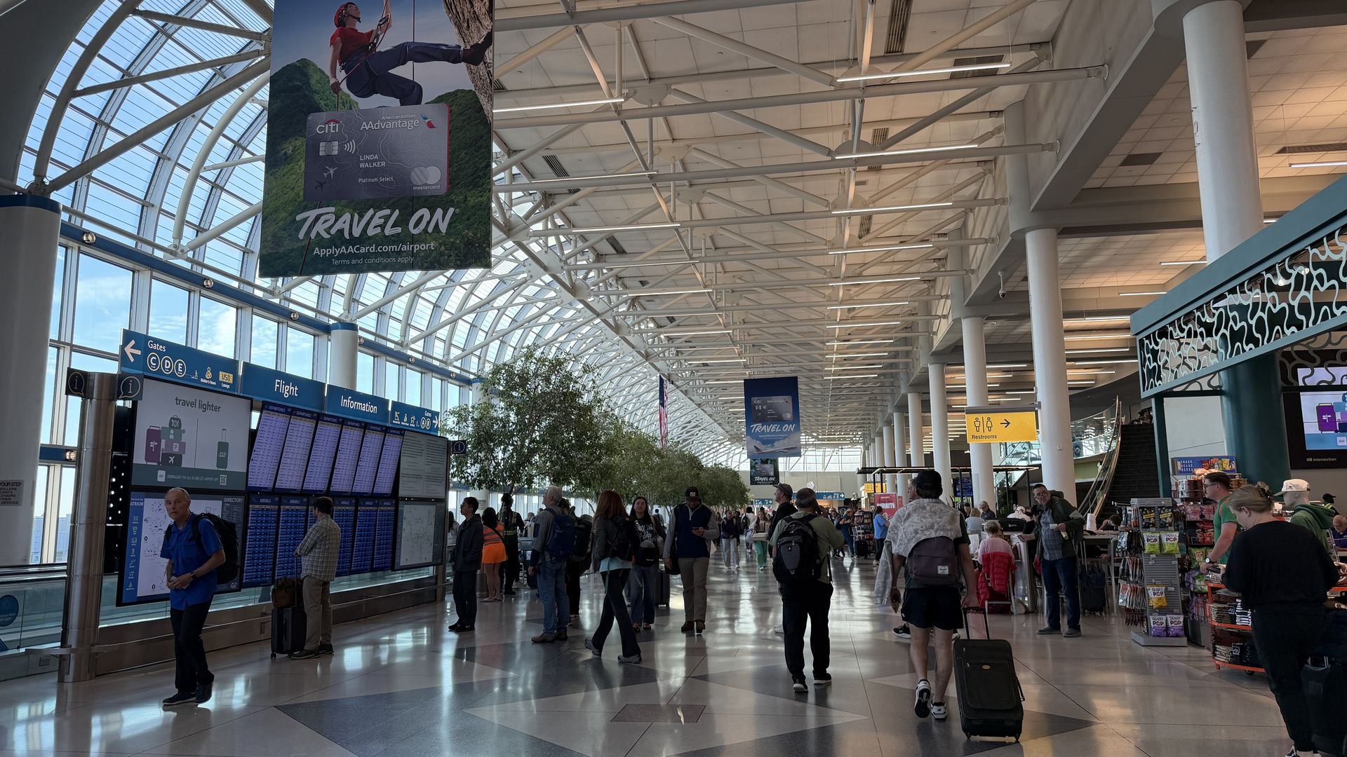 Interior of a busy airport with a glass arched ceiling and white steel beams. Travelers with luggage pass blue gates and flight information boards; a large poster reading "Travel On" hangs overhead.