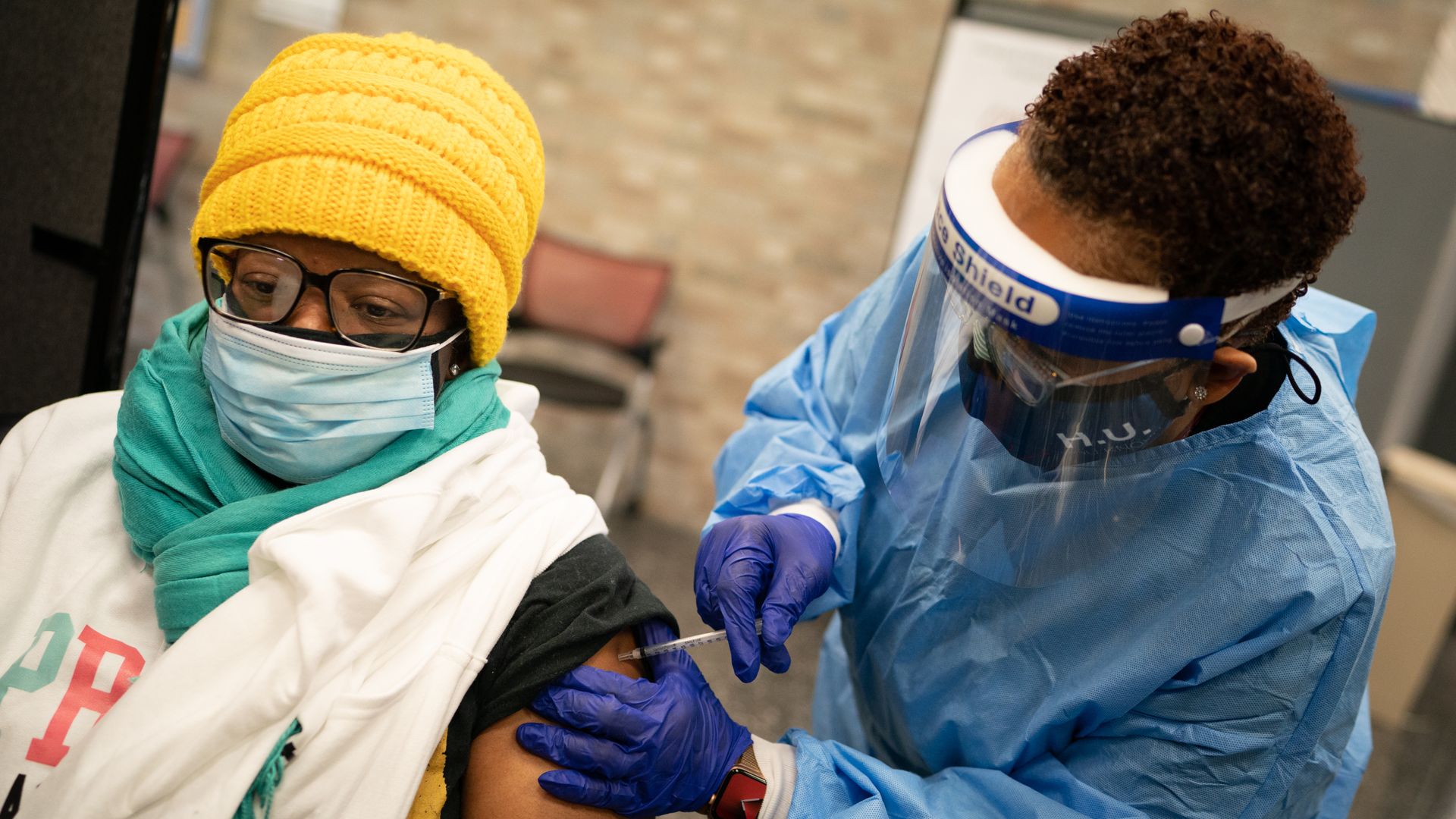 A physician in a blue gown gives a shot to a person in a yellow hat and white face mask