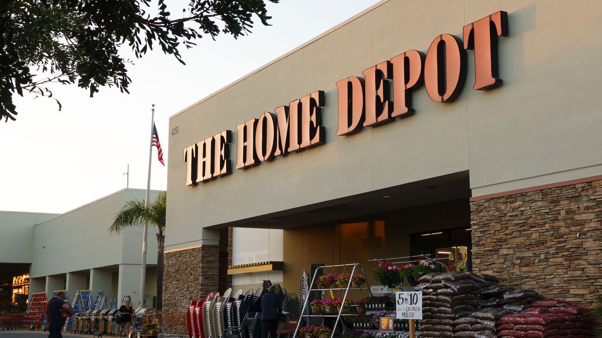 The Home Depot storefront with a large orange sign on a beige wall, an American flag on a pole, overhanging tree branches, and a garden center outside with plants, sacks, and shopping carts.