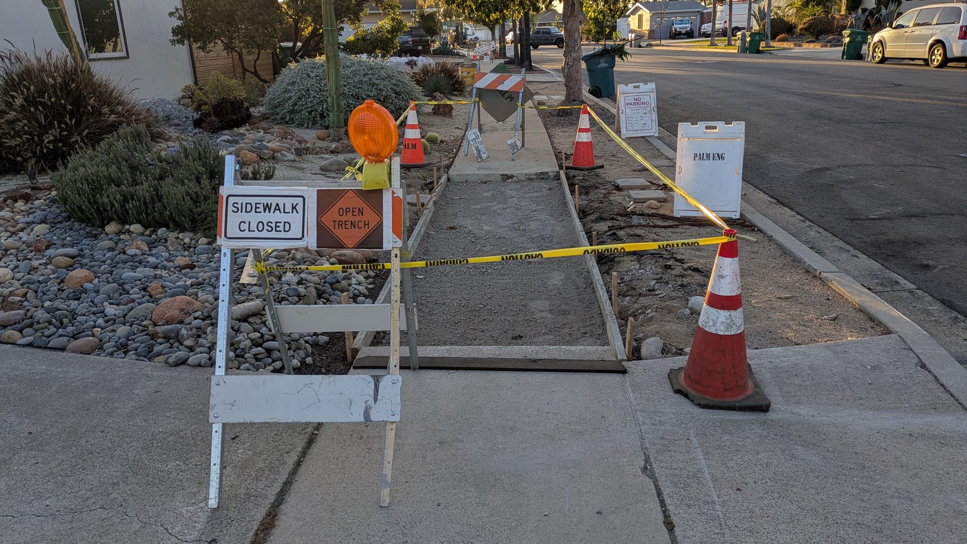Residential street with a sidewalk under construction. Orange cones, yellow caution tape, and a SIDEWALK CLOSED sign near an open trench; rocks and desert plants line the yard at sunset.