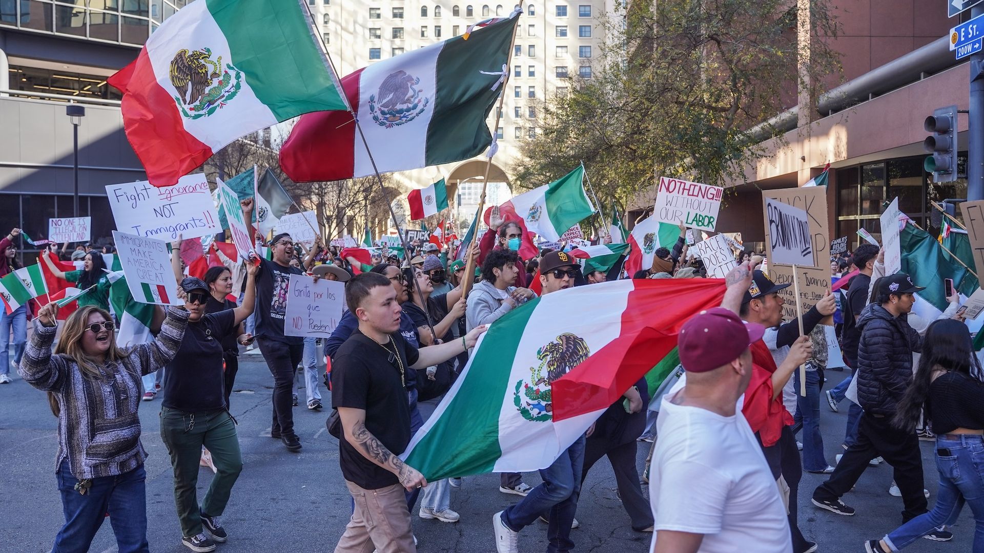 A crowd of people carrying signs and Mexican flags march through city streets during a protest.