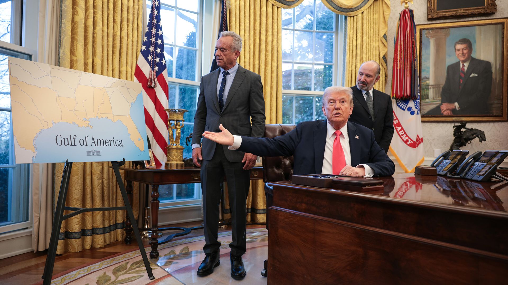 President Trump gestures towards a poster labeled "Gulf of America" while sitting at the Resolute Desk in the Oval Office.