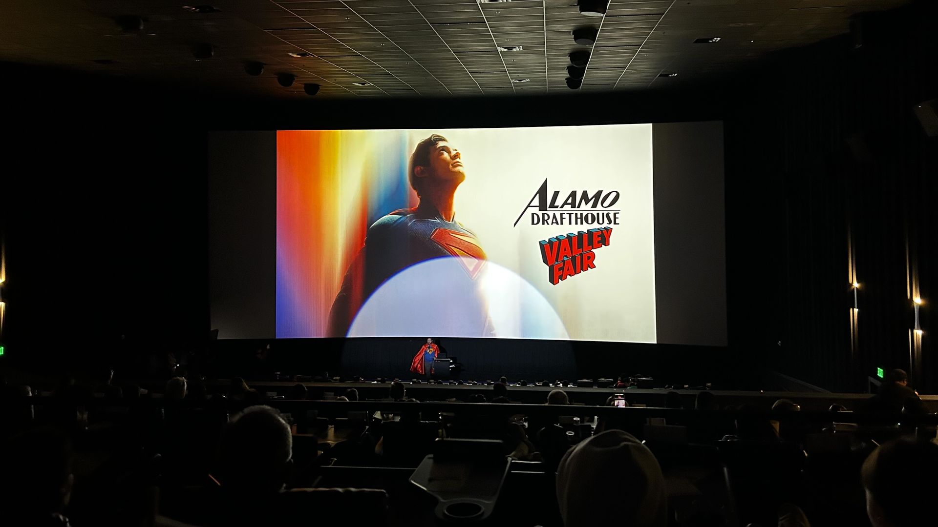 Photo of a large movie theater screen showing the new Superman and the words "Alamo Drafthouse Valley Fair" while a person dressed as Superman speaks 
