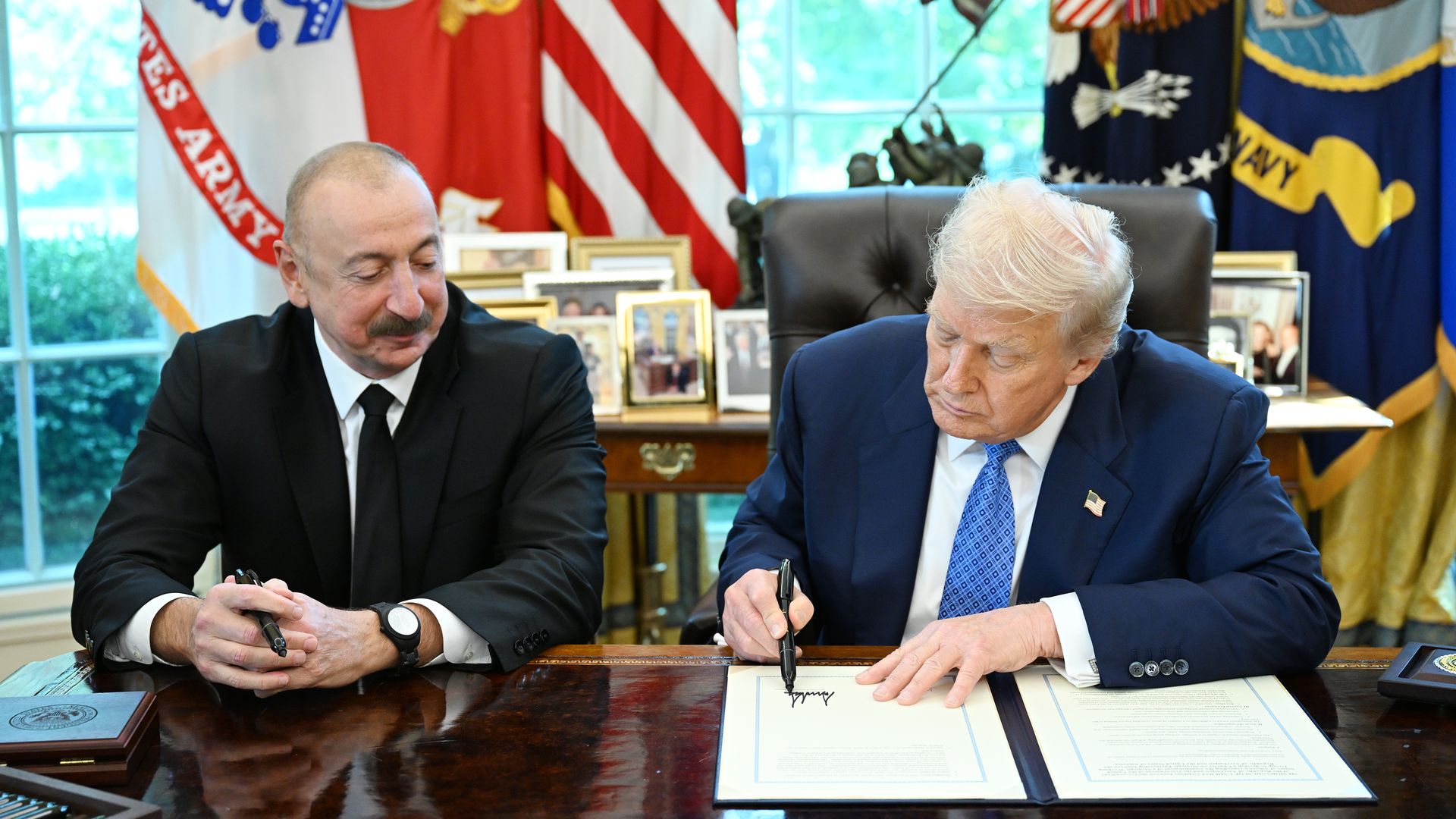 Two men sit at a wooden desk with flags behind them. The man on the left in a black suit watches the man on the right in a blue suit and tie sign a document.