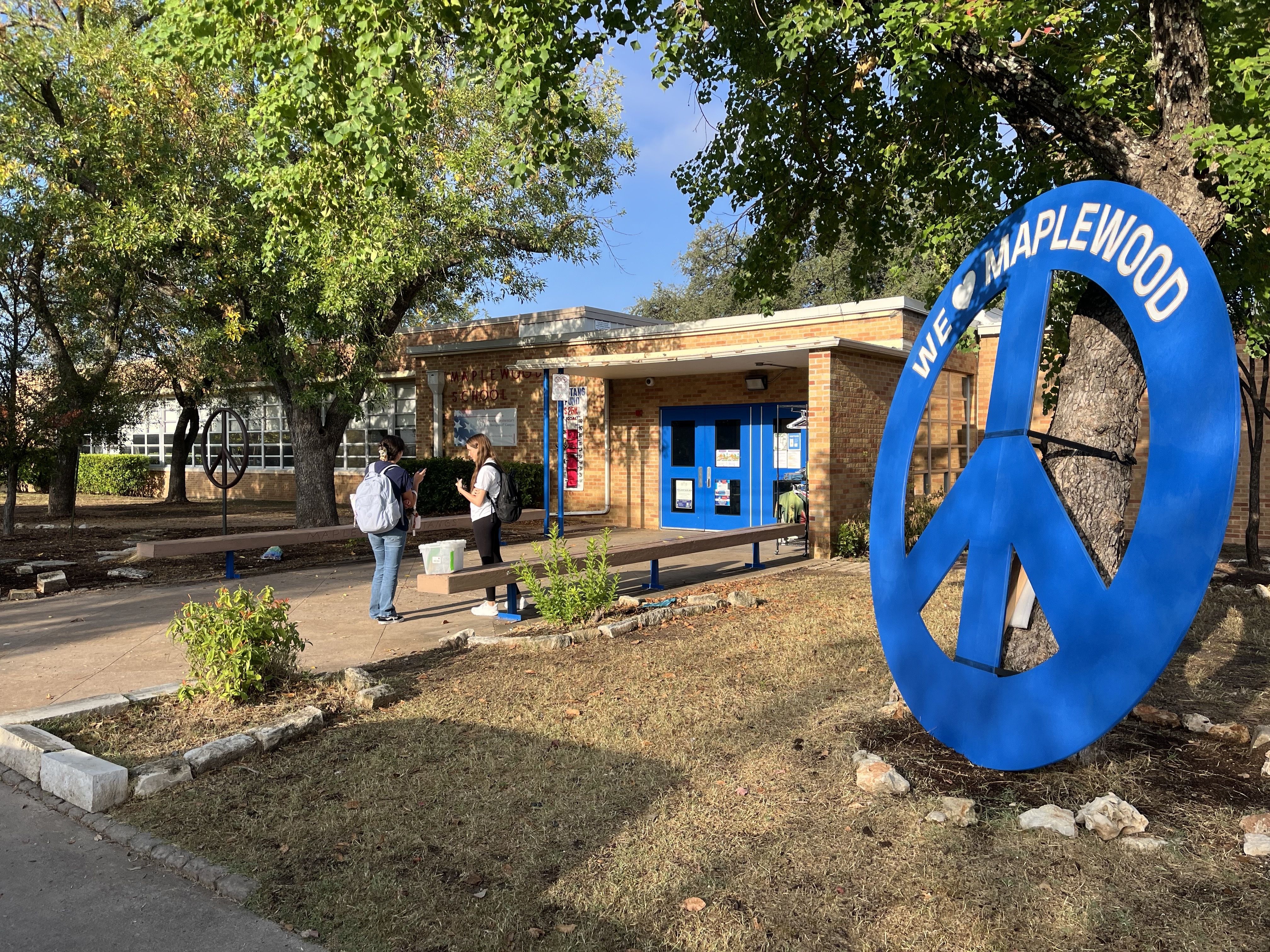 Two people standing outside a brick building with blue double doors, near a large blue peace sign sculpture reading "We ♥ Maplewood" on a sunny day with trees and grass nearby.