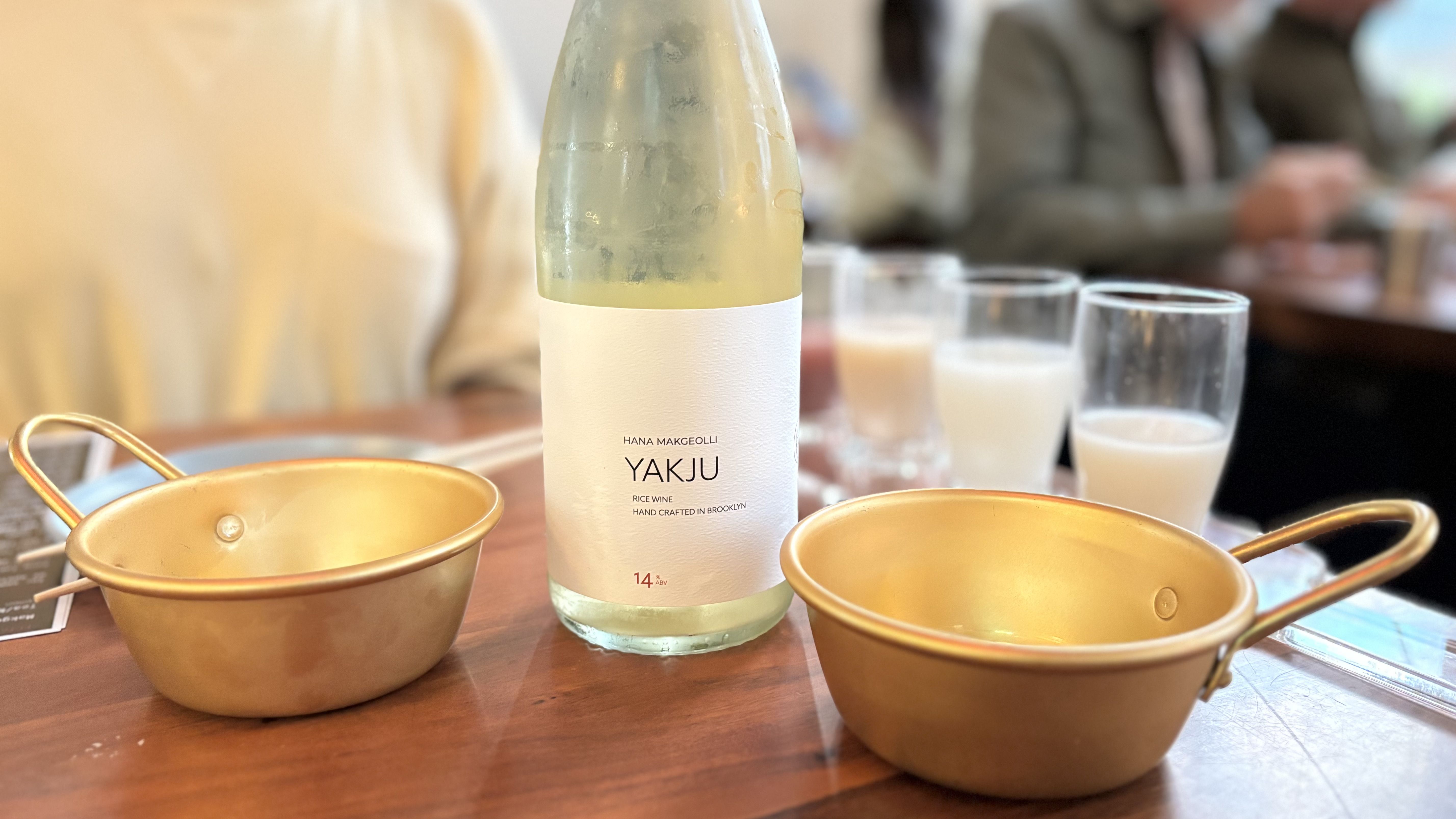 A frosted bottle of Yakju rice wine with a white label sits between two golden metal cups on a wooden table, with three small glasses of milky white rice wine in the background.