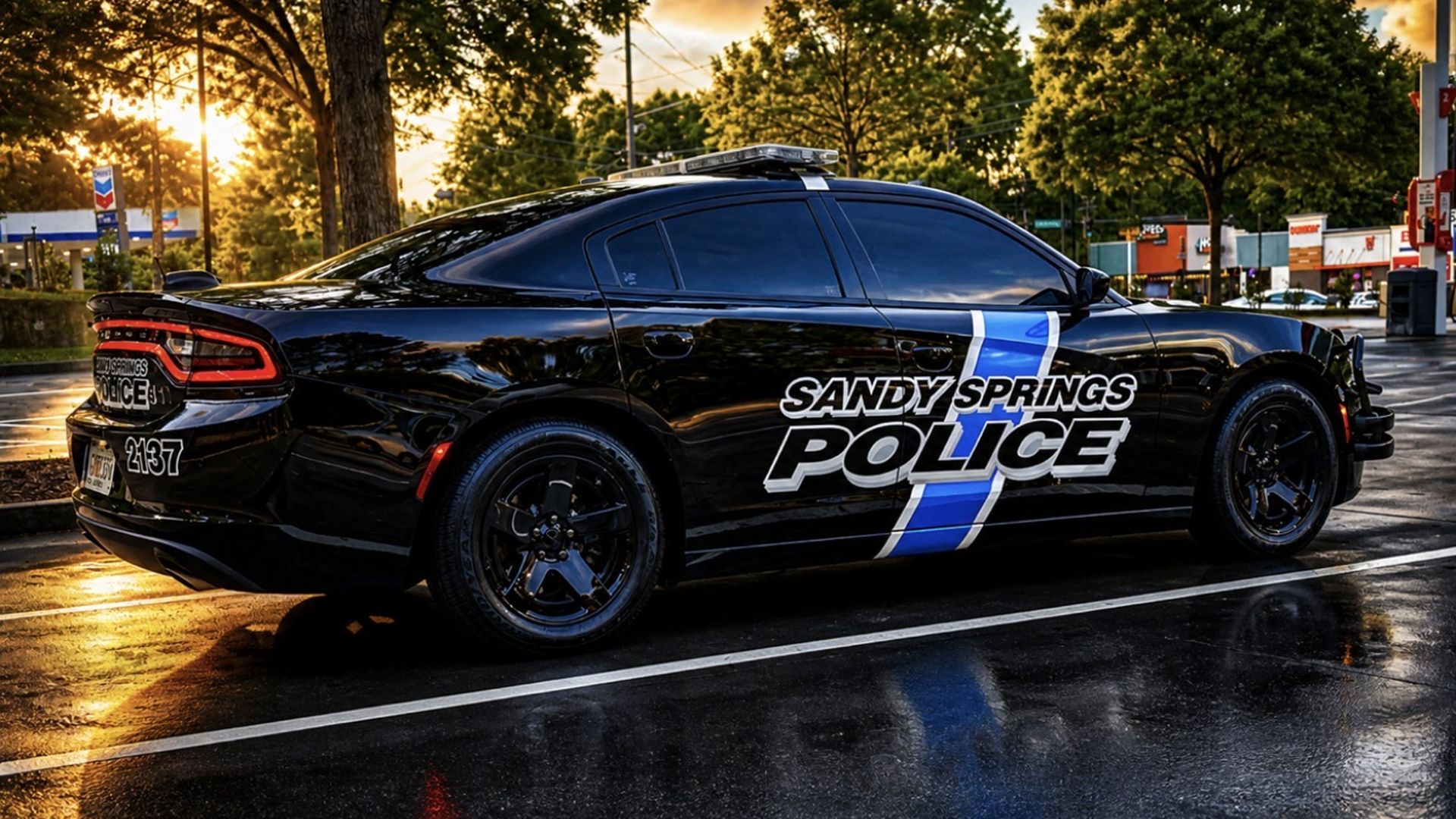 A glossy black Sandy Springs Police Dodge Charger parked in a wet parking lot at sunset. The side shows a bold white and blue "SANDY SPRINGS POLICE" logo with a diagonal blue stripe.