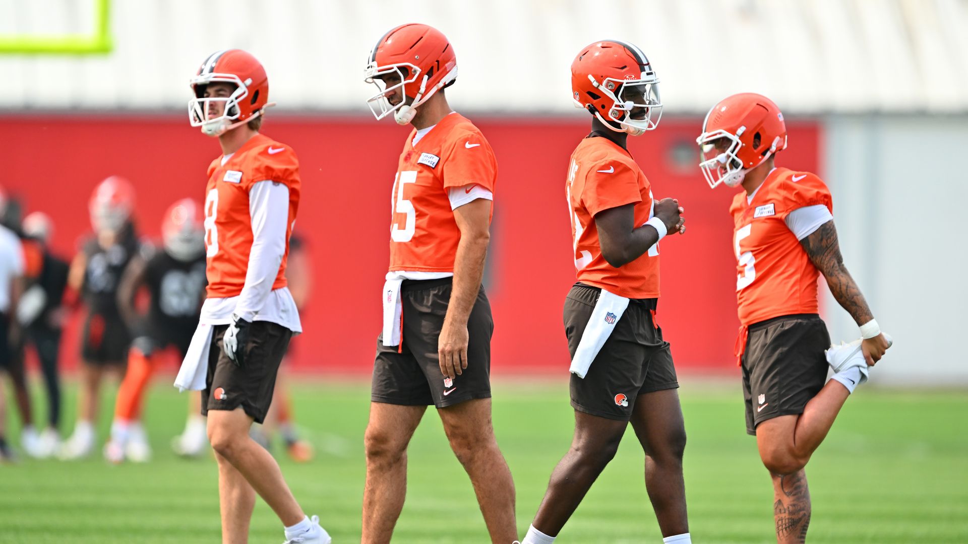 Four football players in orange jerseys and helmets standing on a green field, two stretching and two standing relaxed during practice session.