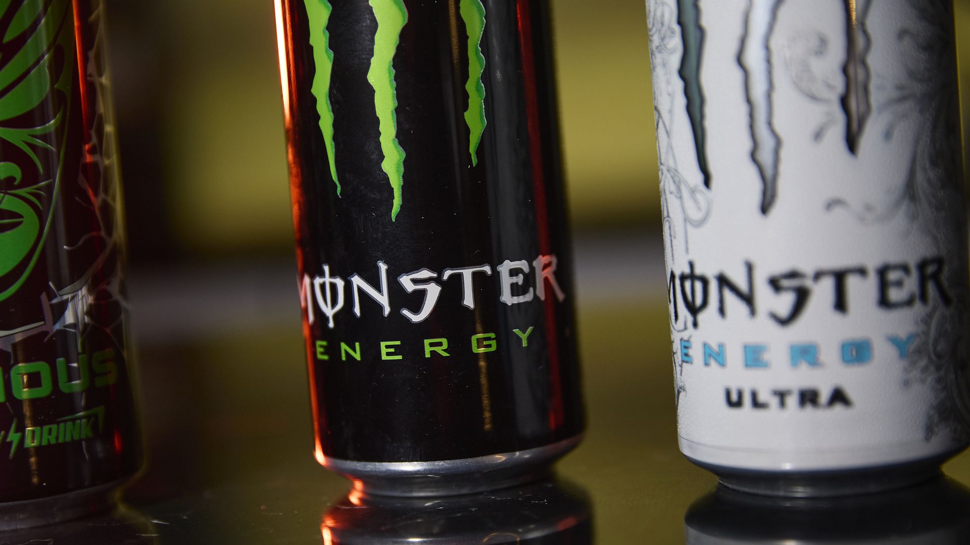 Close-up of two Monster Energy cans on a glossy surface: a black can with the green claw logo center, a white Ultra can to the right, and partial red/green can on the left, with reflections.