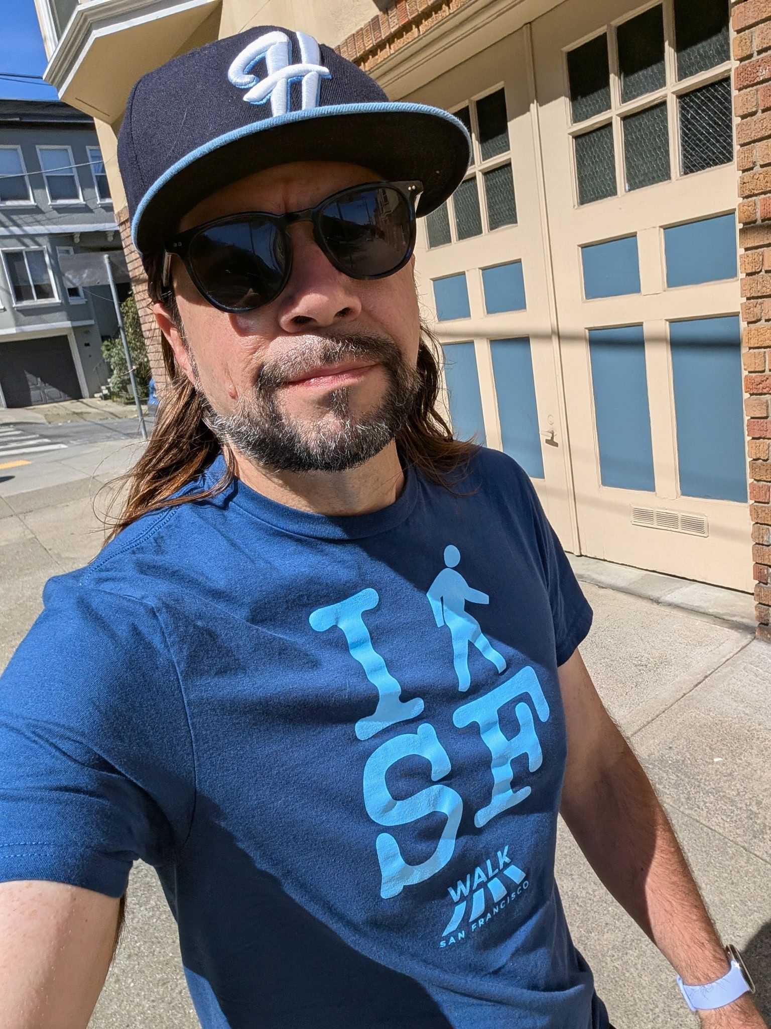 Man wearing sunglasses and a navy cap with a light blue logo, blue t-shirt with a walking figure and WALK; standing on a sunny sidewalk outside a brick building with a beige garage door.