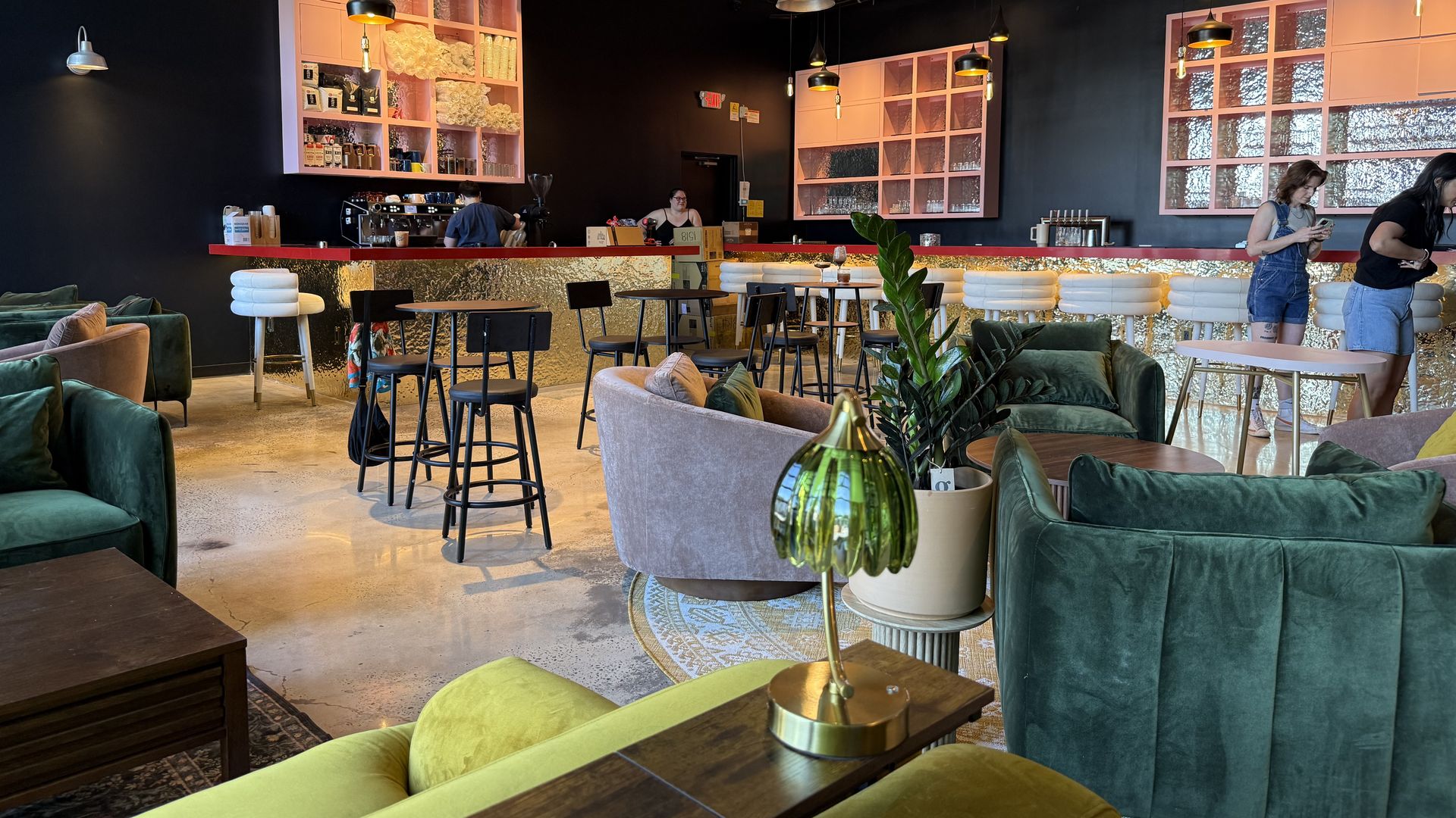 A stylish cafe interior featuring a gold-textured bar with pink shelving, black walls, and exposed ceiling pipes; green velvet seating, and a few customers near the bar.